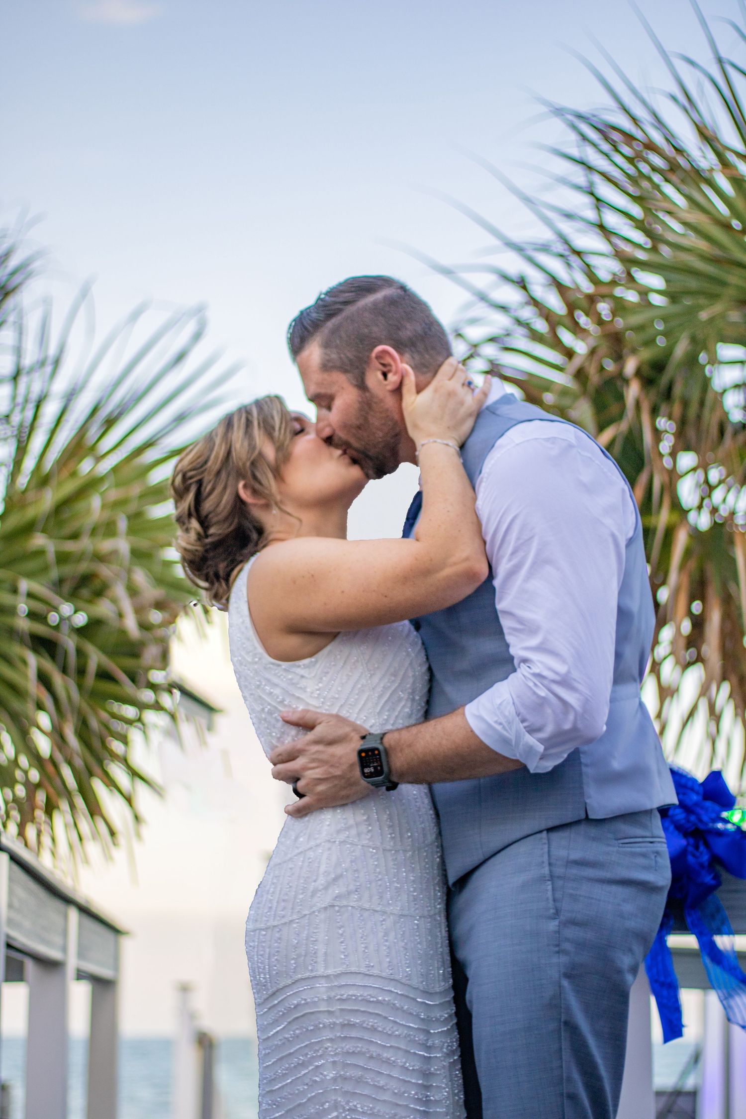 A romantic kiss between newlyweds with palm trees and ocean in the background.