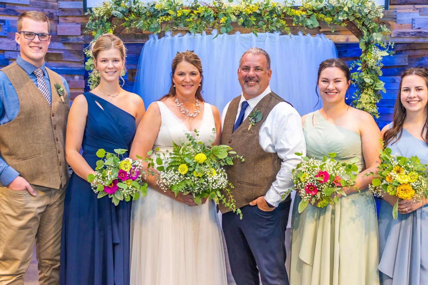 A wedding party poses together under a floral arch with blue drapery backdrop.
