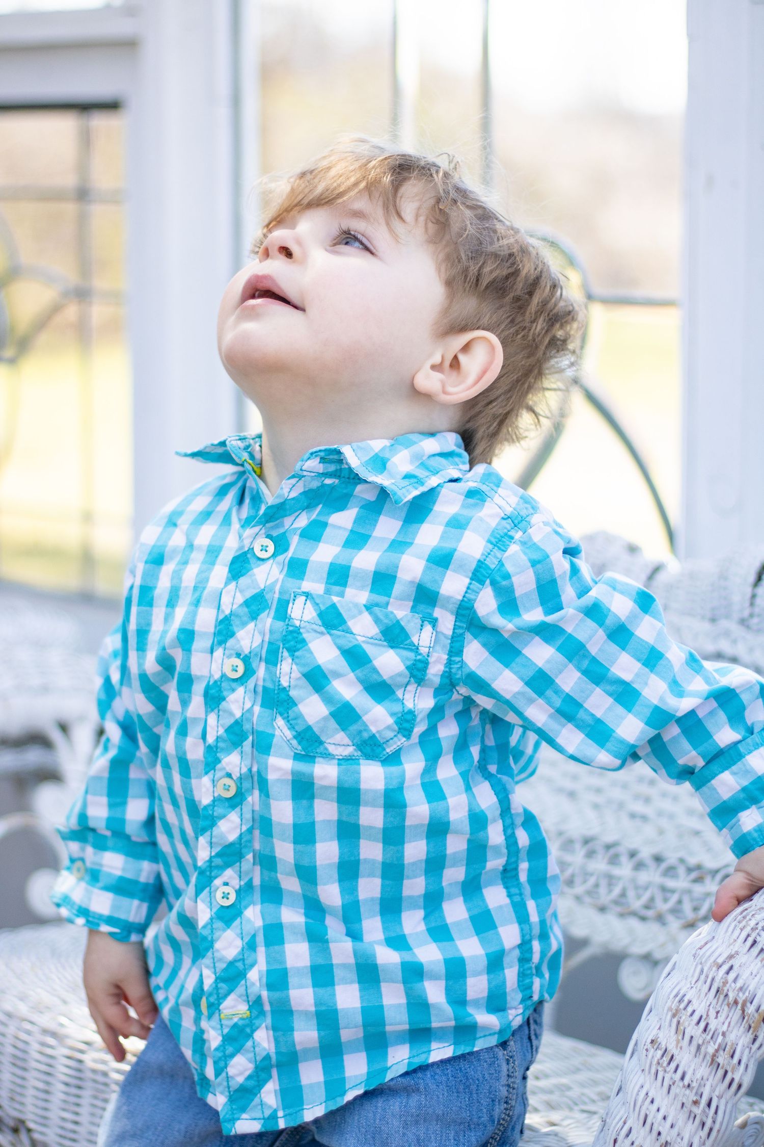 A toddler in blue gingham shirt looks up while standing near wicker furniture.