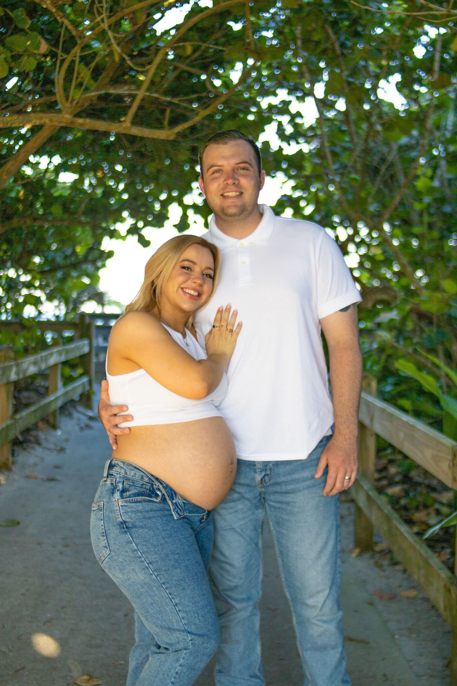 A happy expecting couple poses together on a wooden walkway surrounded by greenery.