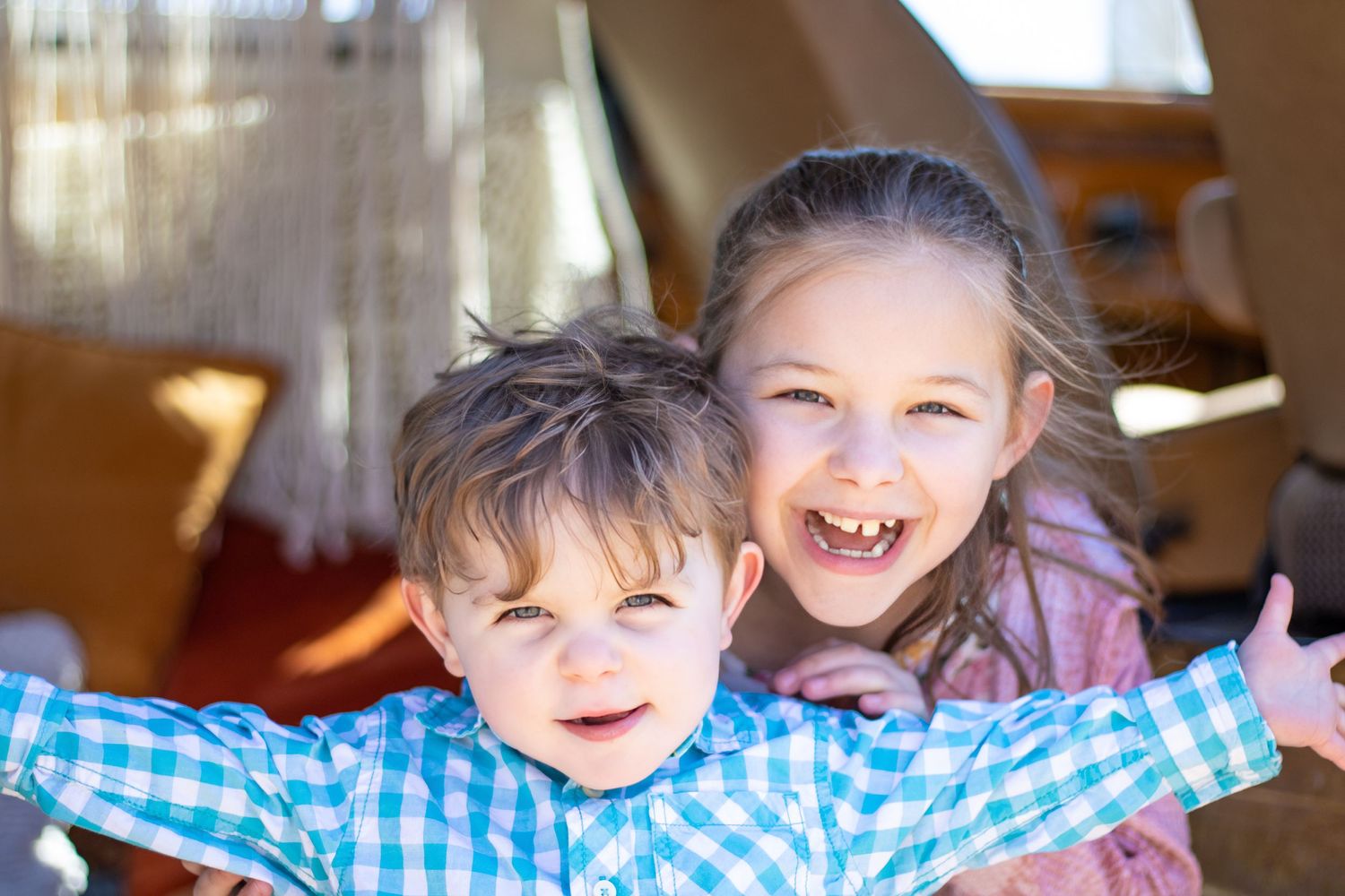 Two playful children laughing and smiling in front of a bright window.