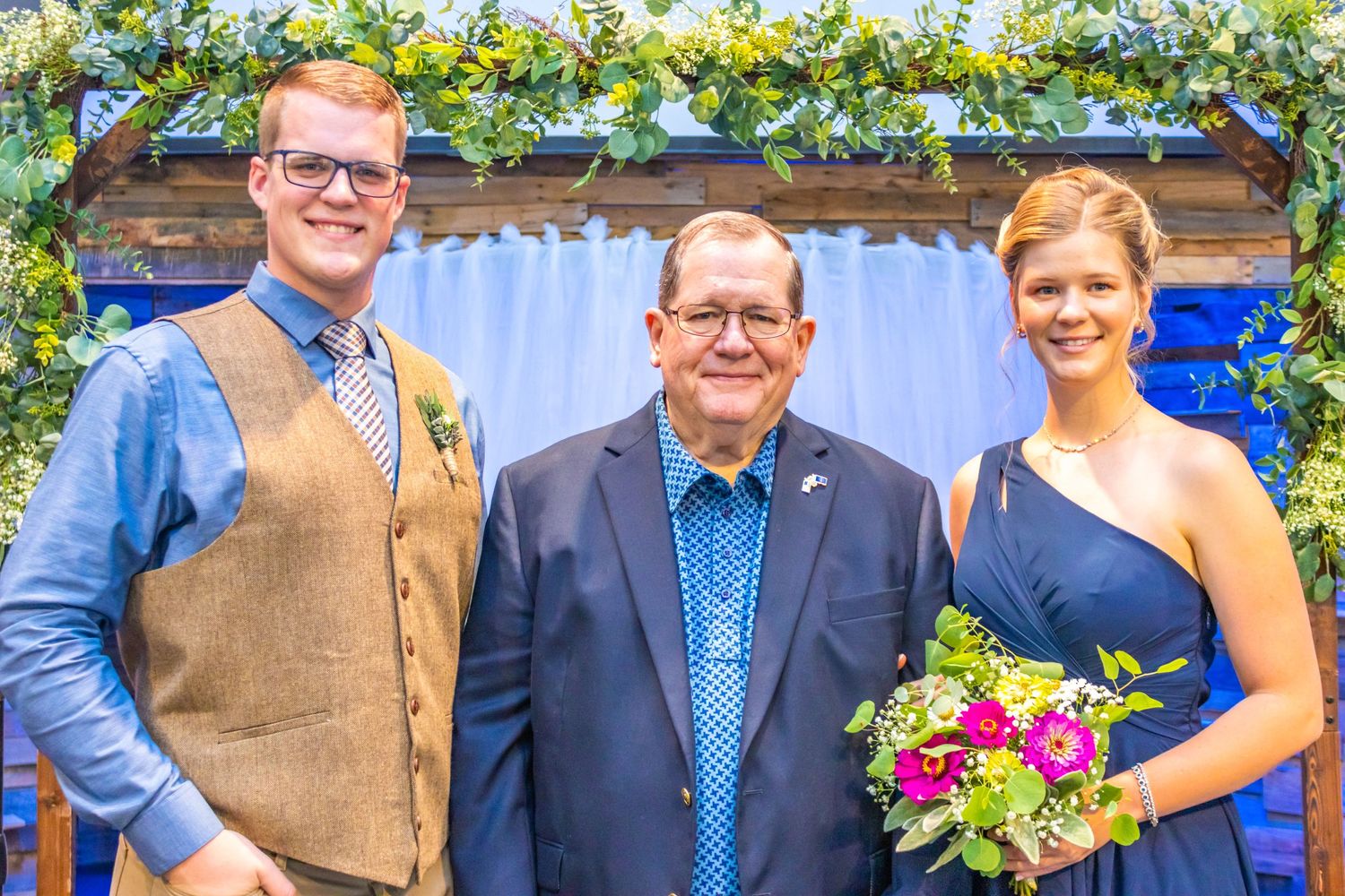 Group poses under floral decorated arch with blue backdrop at formal event.
