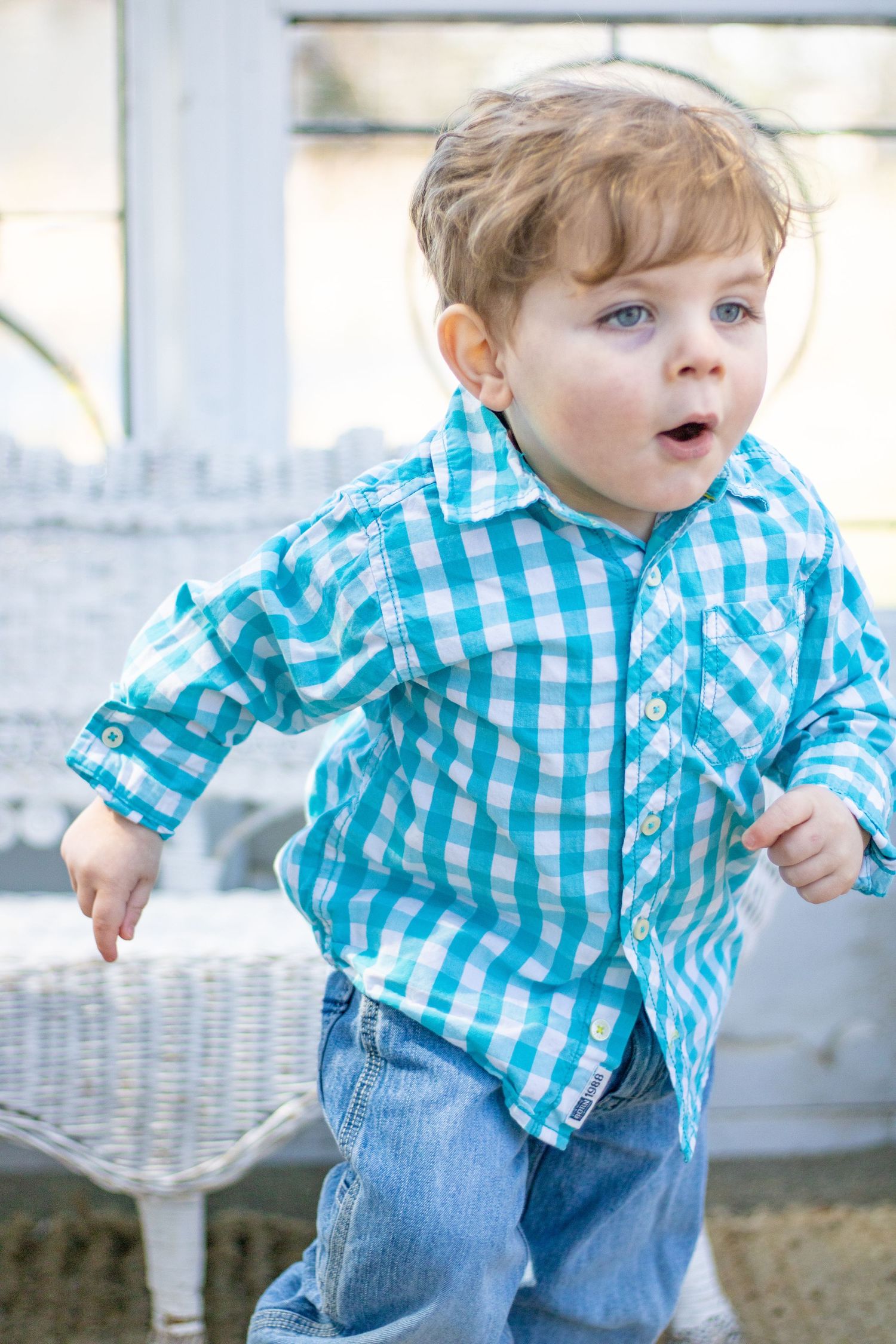 A toddler in blue checkered shirt and jeans standing by white wicker furniture.