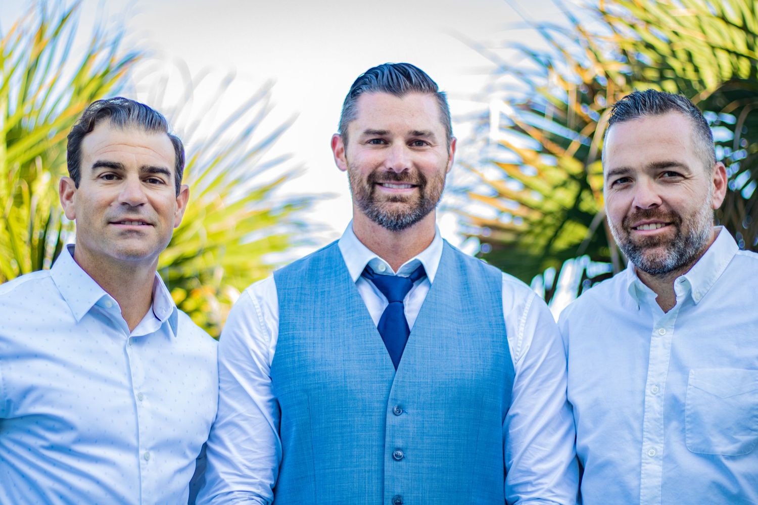 Three men in formal attire pose against a tropical palm tree background.