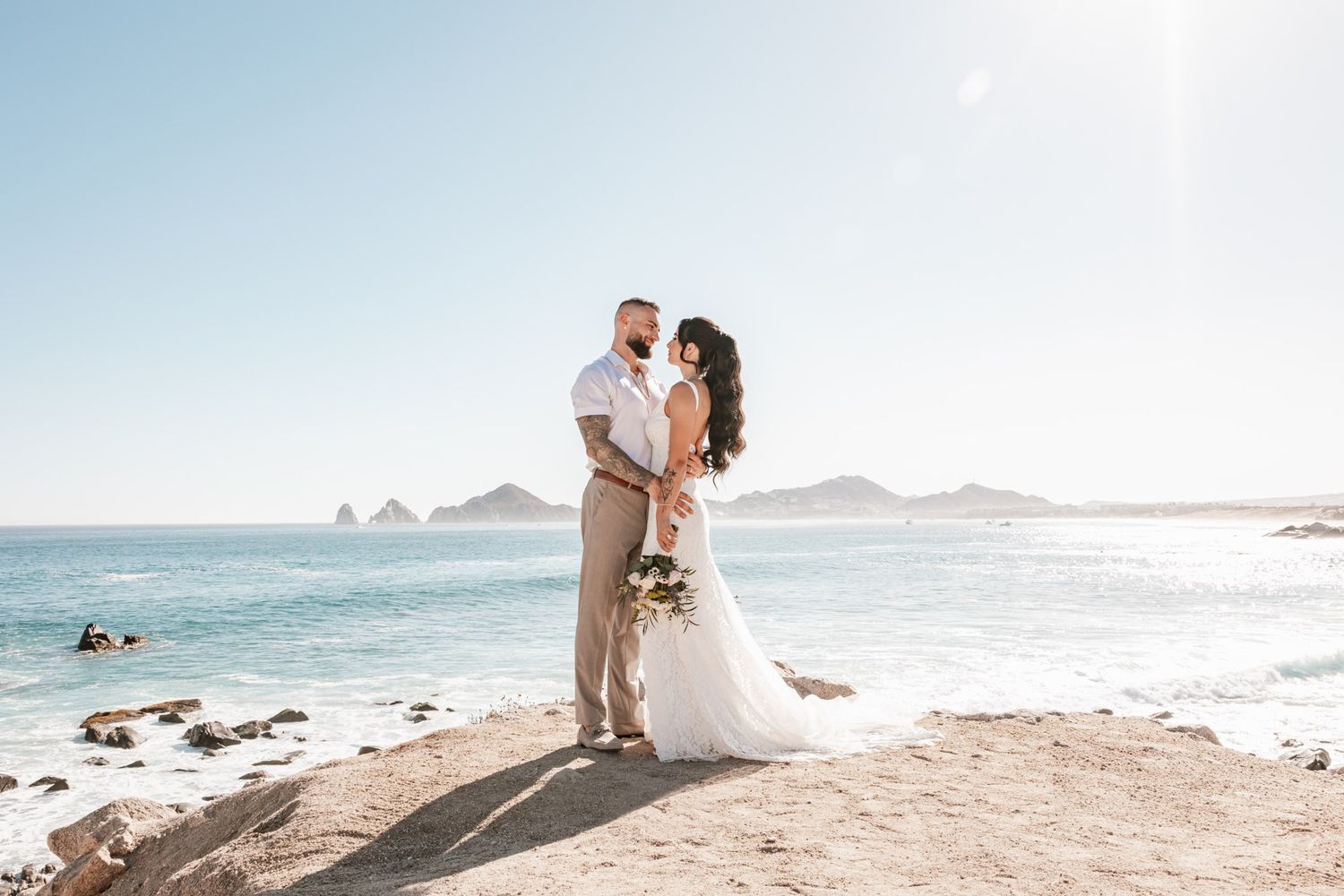 A couple in wedding attire embraces on a scenic beach with rocky coastline and blue ocean waters in the background.