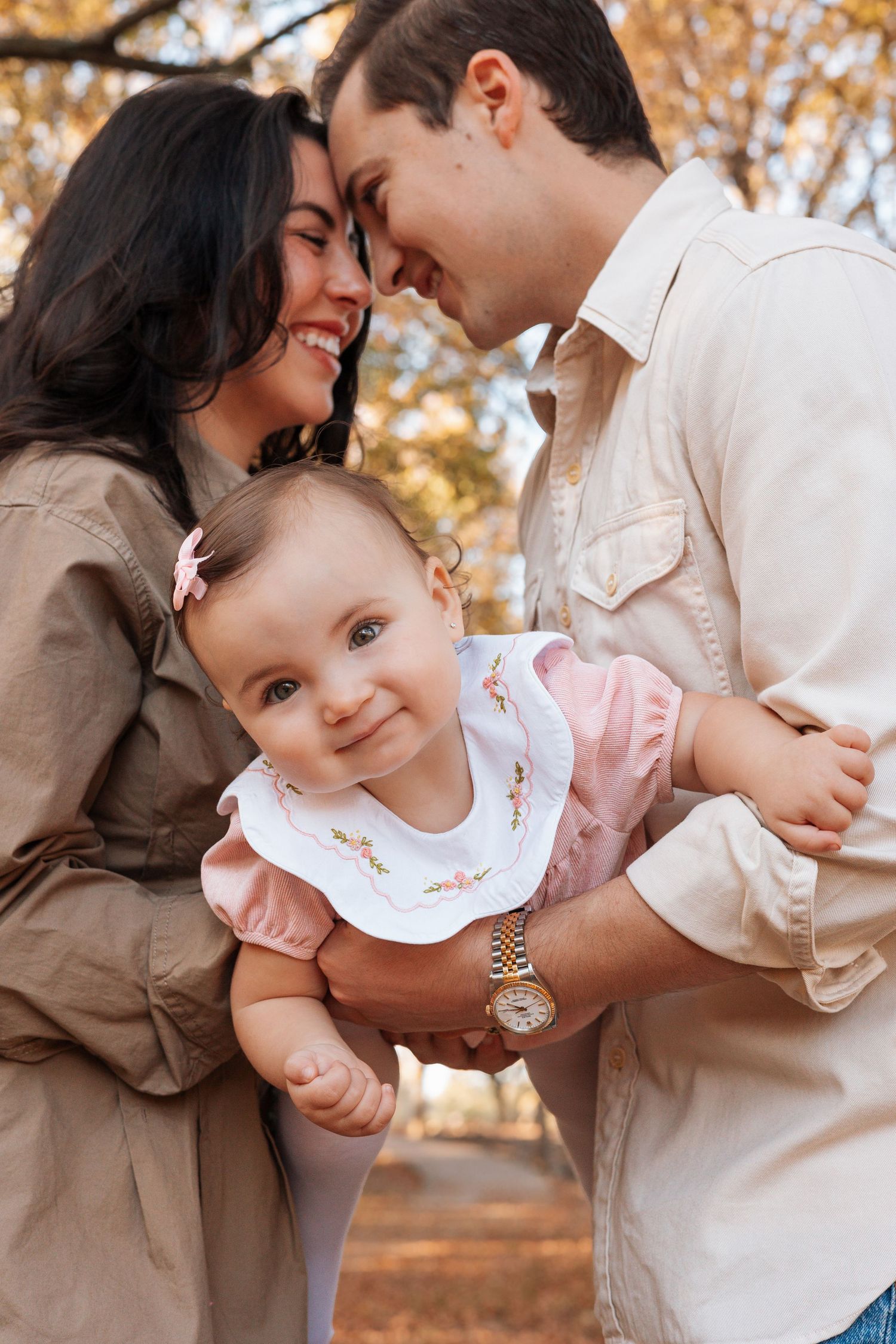 A couple shares tender moments with their baby outdoors during autumn with golden leaves in the background.