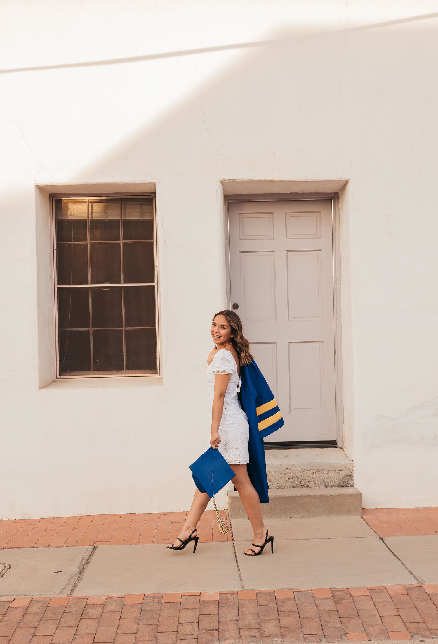 Young woman in white dress holding blue graduation cap and gown, posing outdoors near a white building.