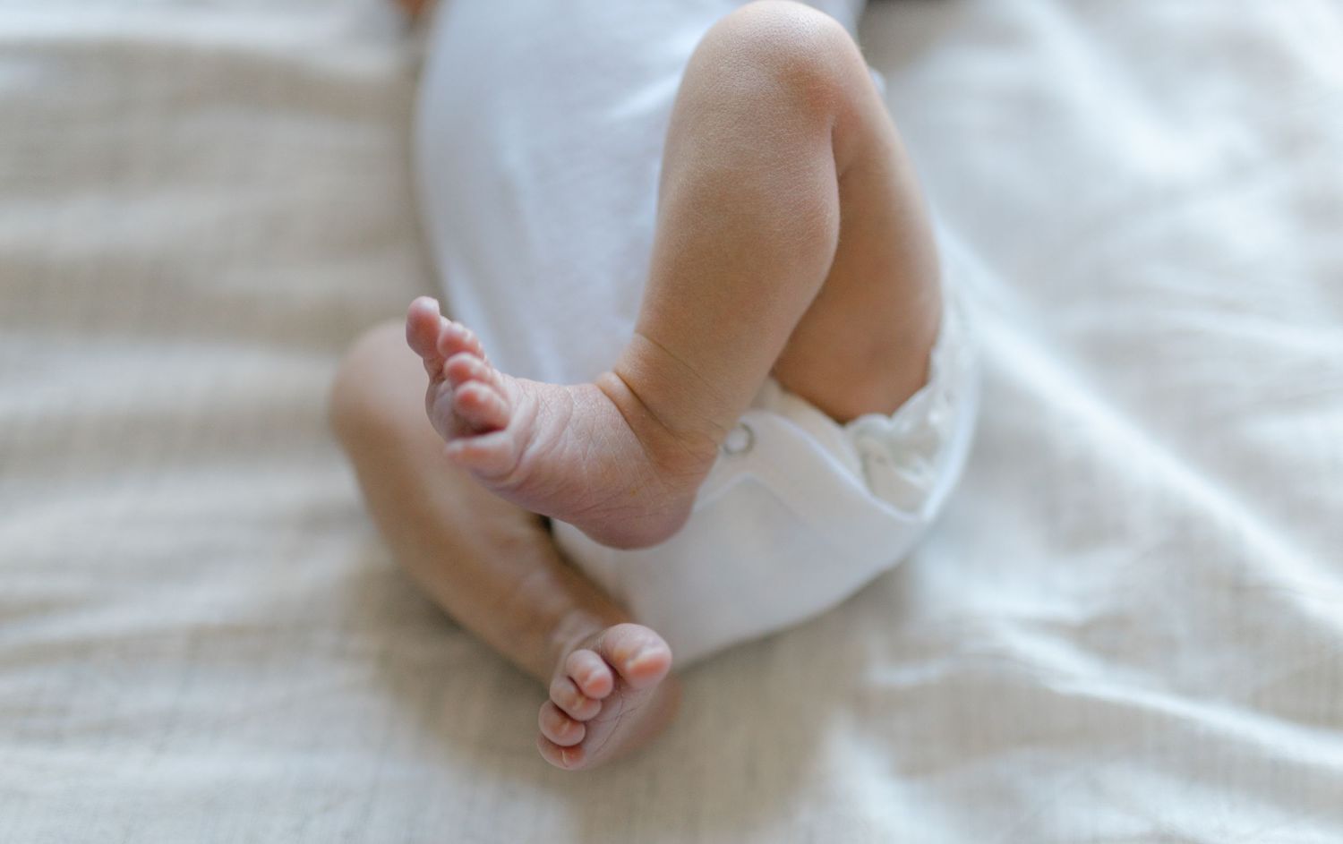 Tiny bare feet and chubby legs of a baby resting on soft white bedding.