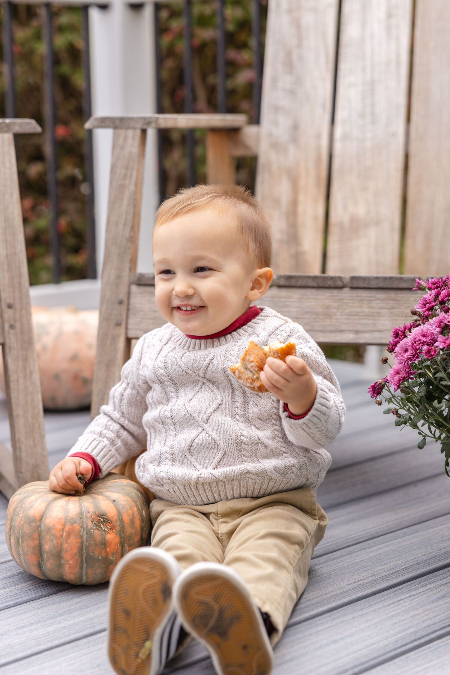Baby in white sweater sits on wooden deck with pumpkins and pink flowers in autumn setting.