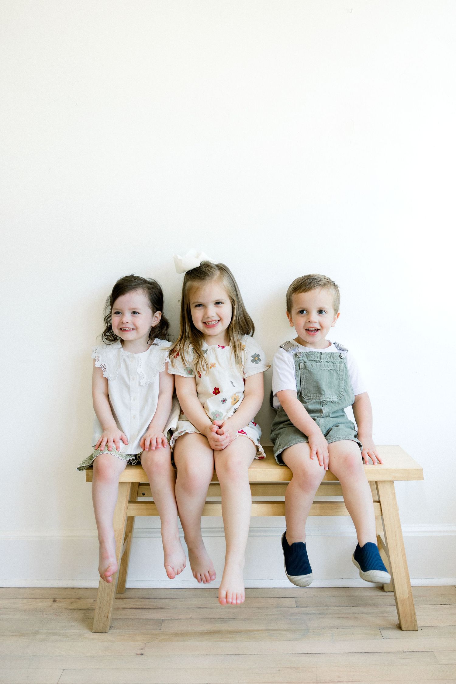 Three young children sit together on a wooden bench against a white wall, wearing casual summer outfits.