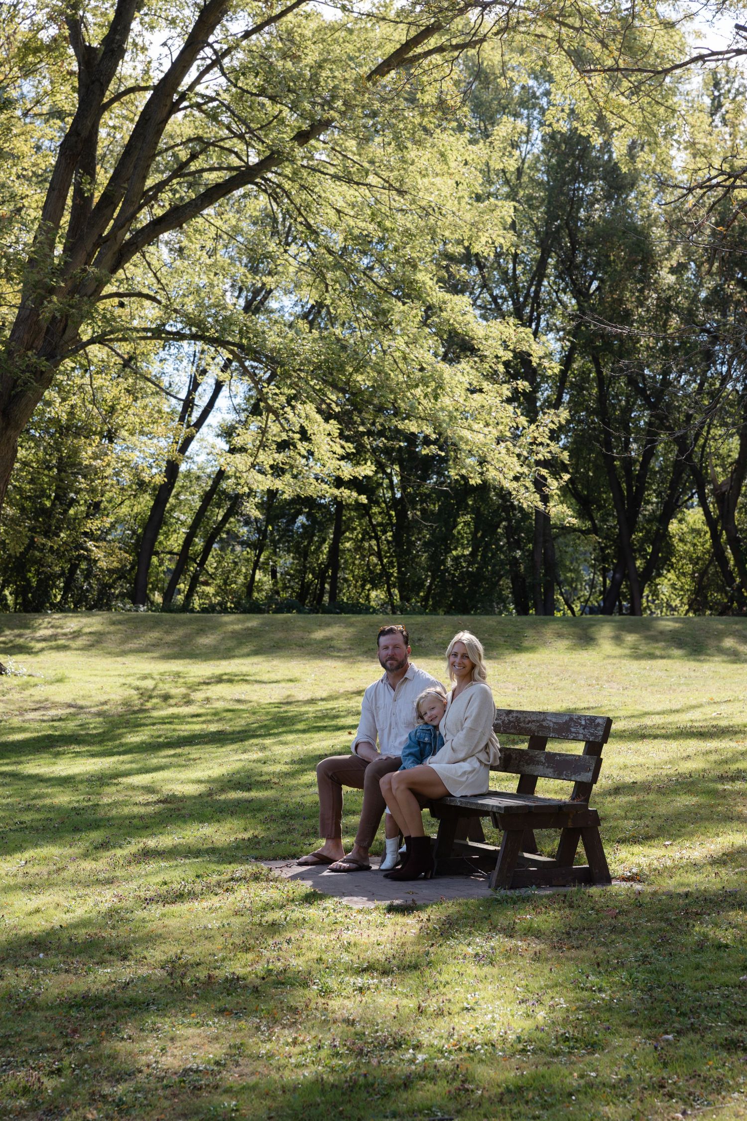 A couple relaxes together on a wooden bench in a sunny park surrounded by tall green trees on a peaceful afternoon.