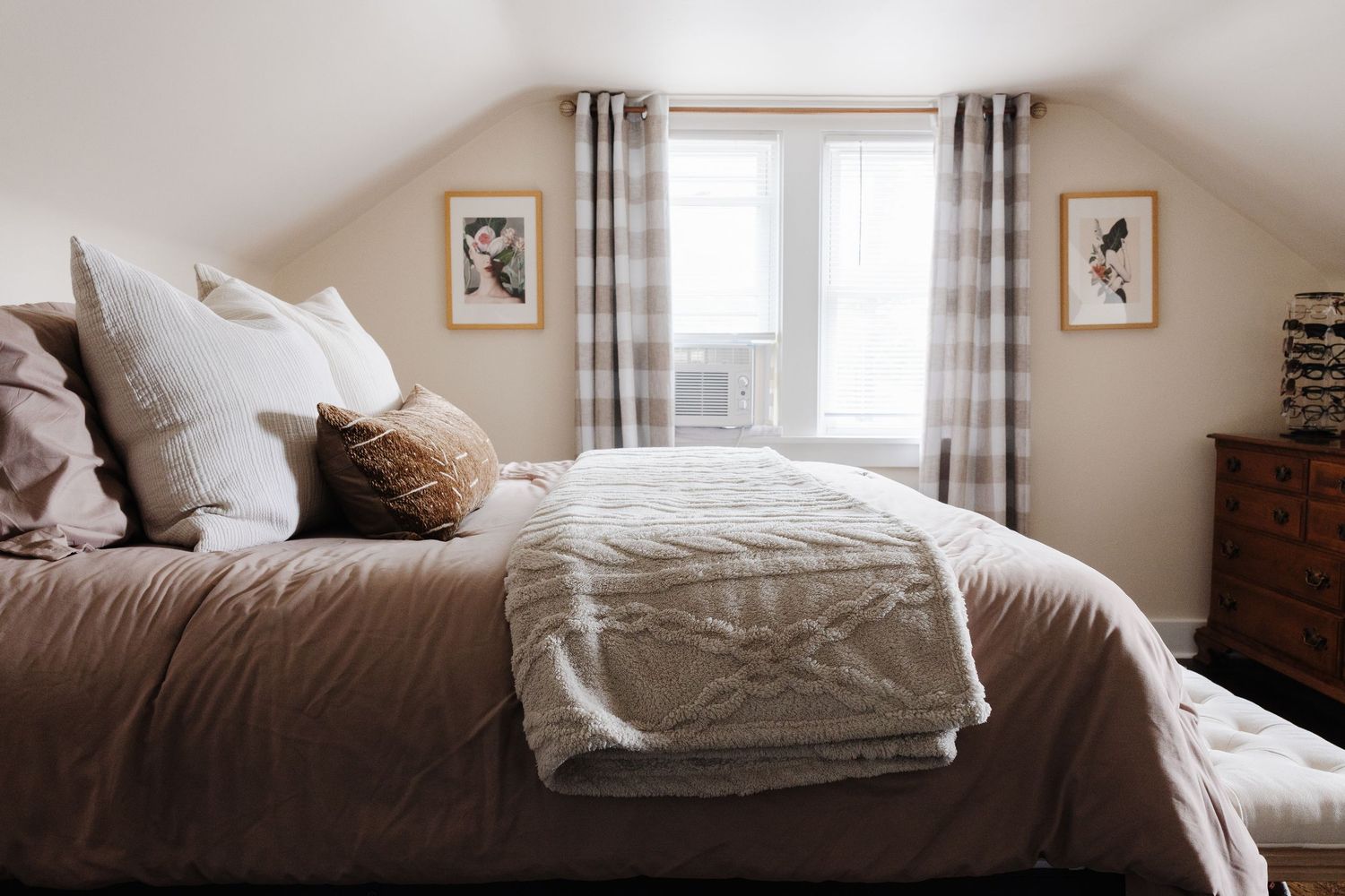 A cozy bedroom interior with white bedding, wooden furniture, and natural light streaming through the window.