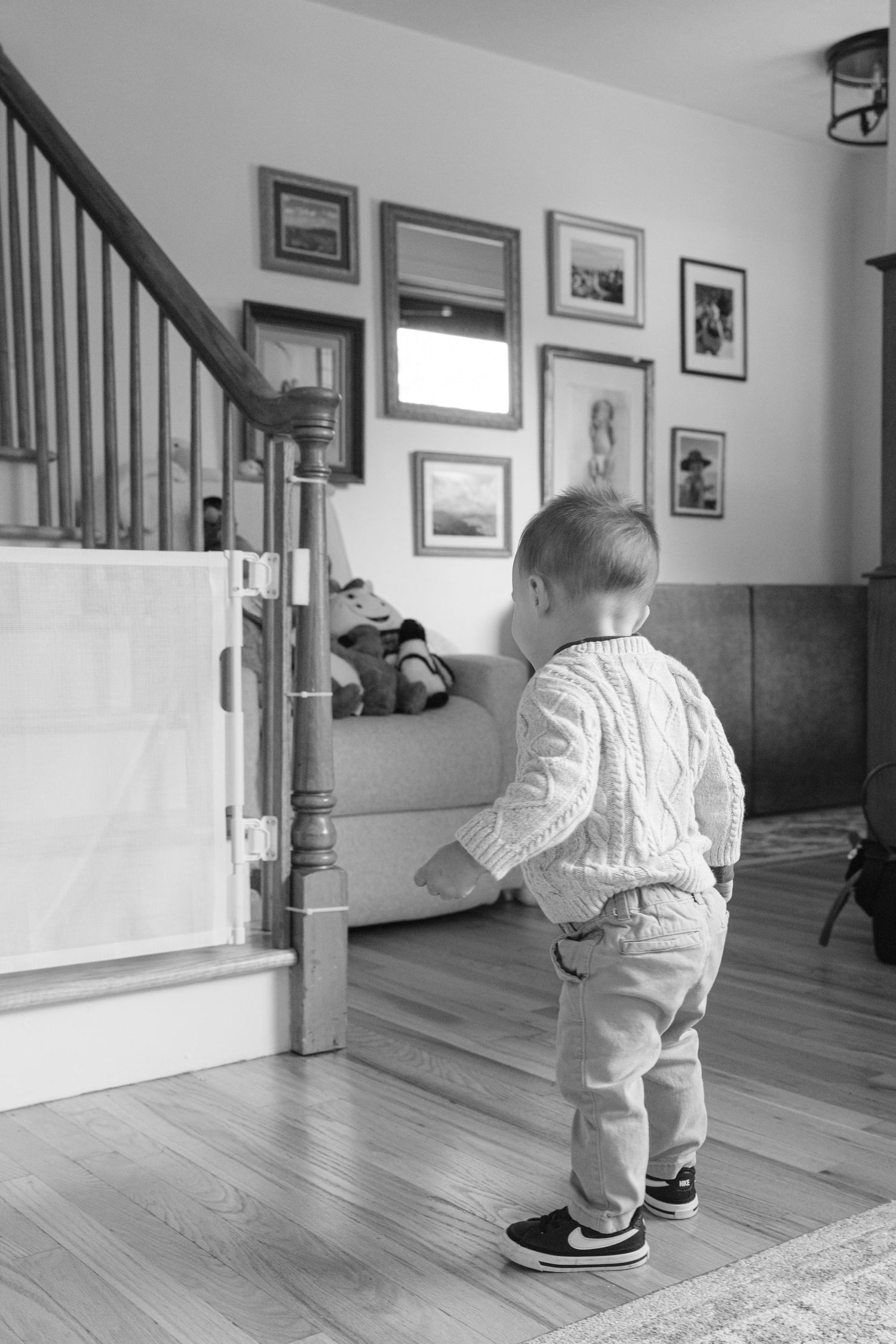 A toddler in a cable knit sweater stands near a baby gate at the bottom of stairs with framed photos on the wall.
