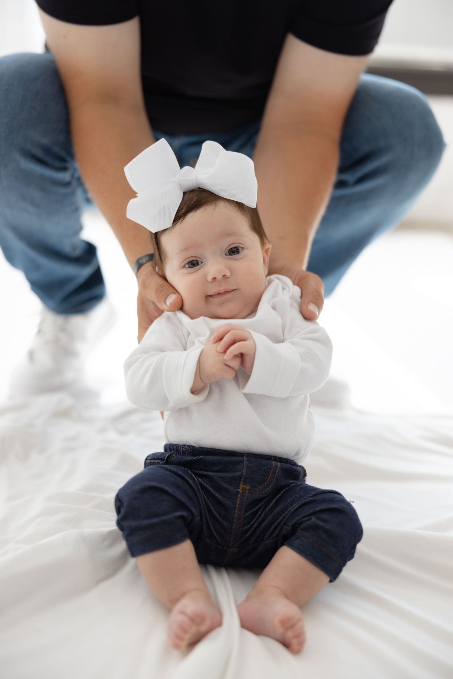 Baby wearing white bow wearing navy pants and white top laying on white bedding with adult hands supporting.