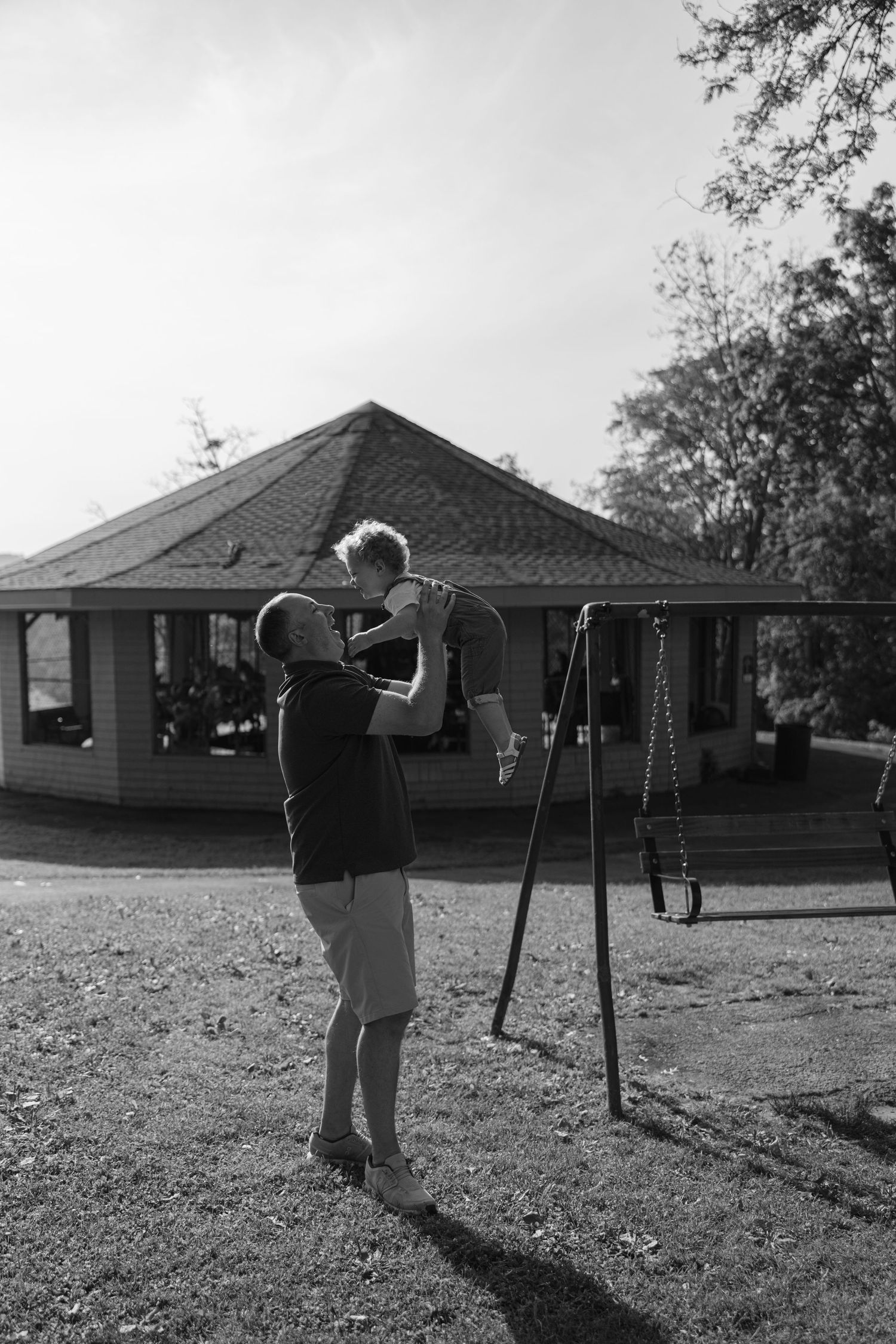 A black and white photo shows someone holding a small child near a swing set outside a suburban house.