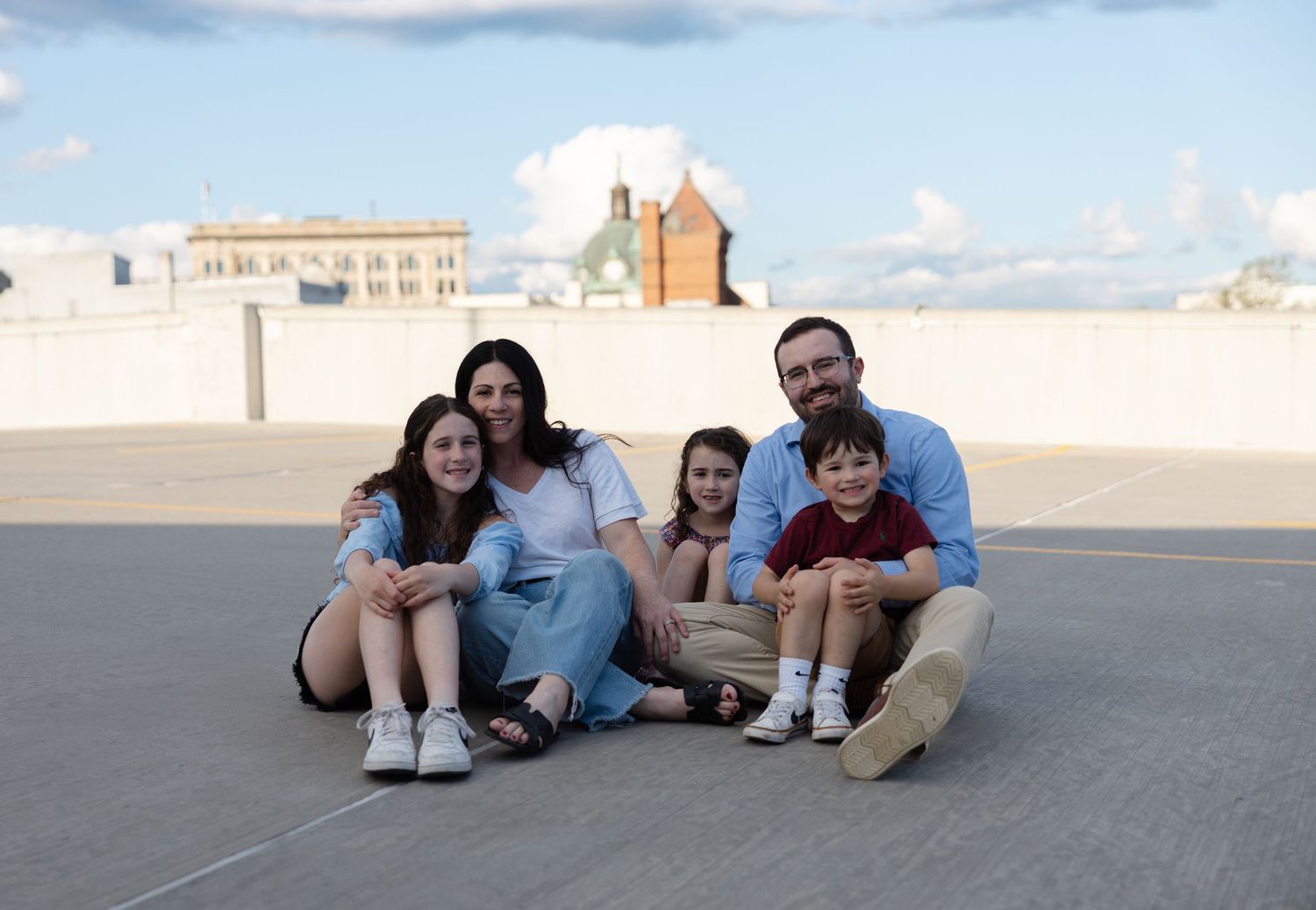 A family sits together on an outdoor rooftop or terrace with buildings and blue sky visible in the background.
