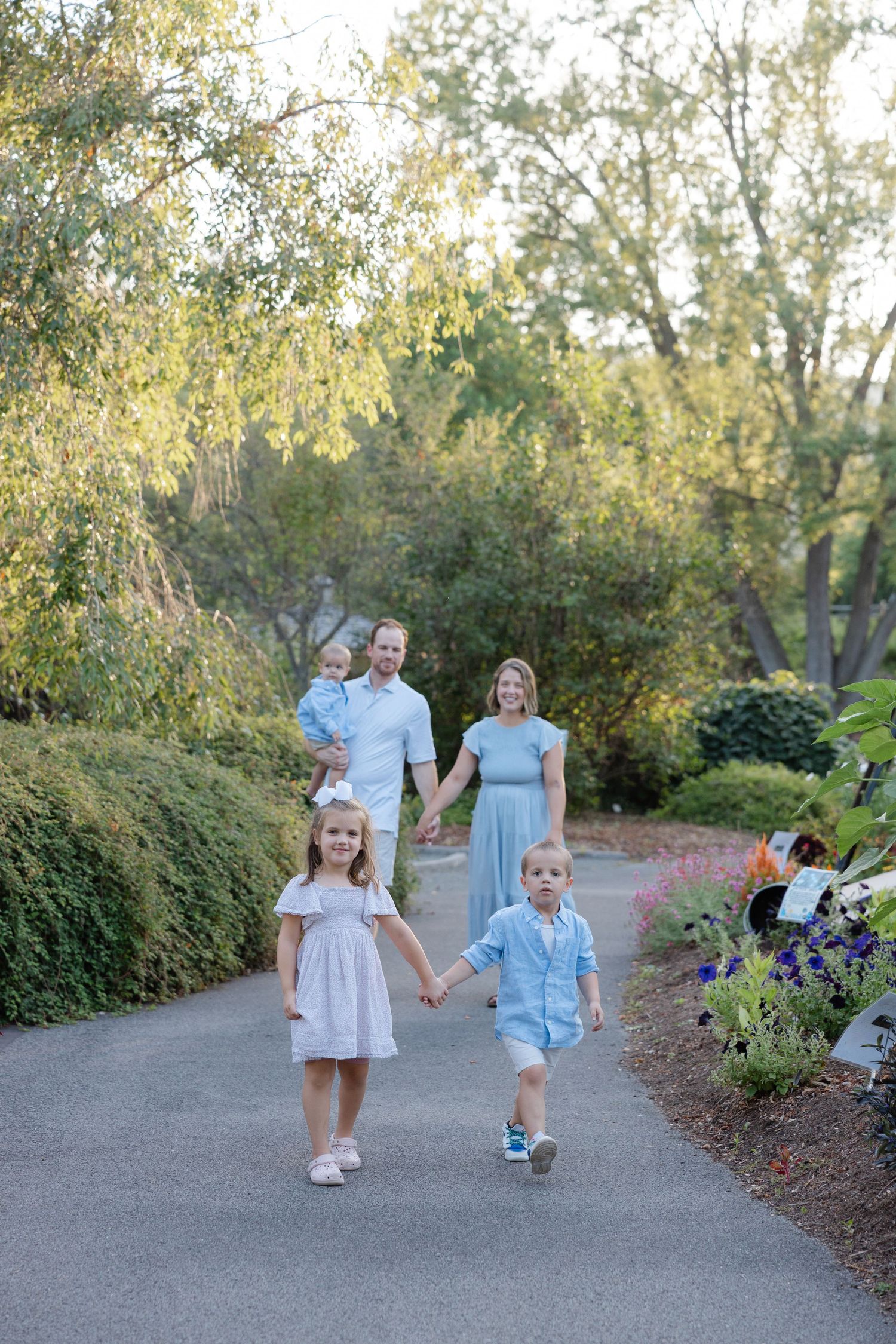 A group of children in light blue and white outfits walk together on a garden path surrounded by lush greenery.