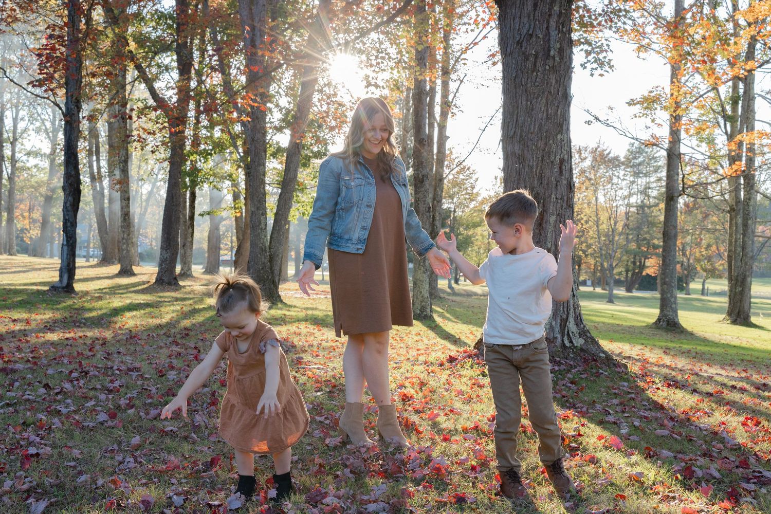 People enjoying a sunny autumn day in a park with golden leaves scattered on the ground as sunlight filters through trees.