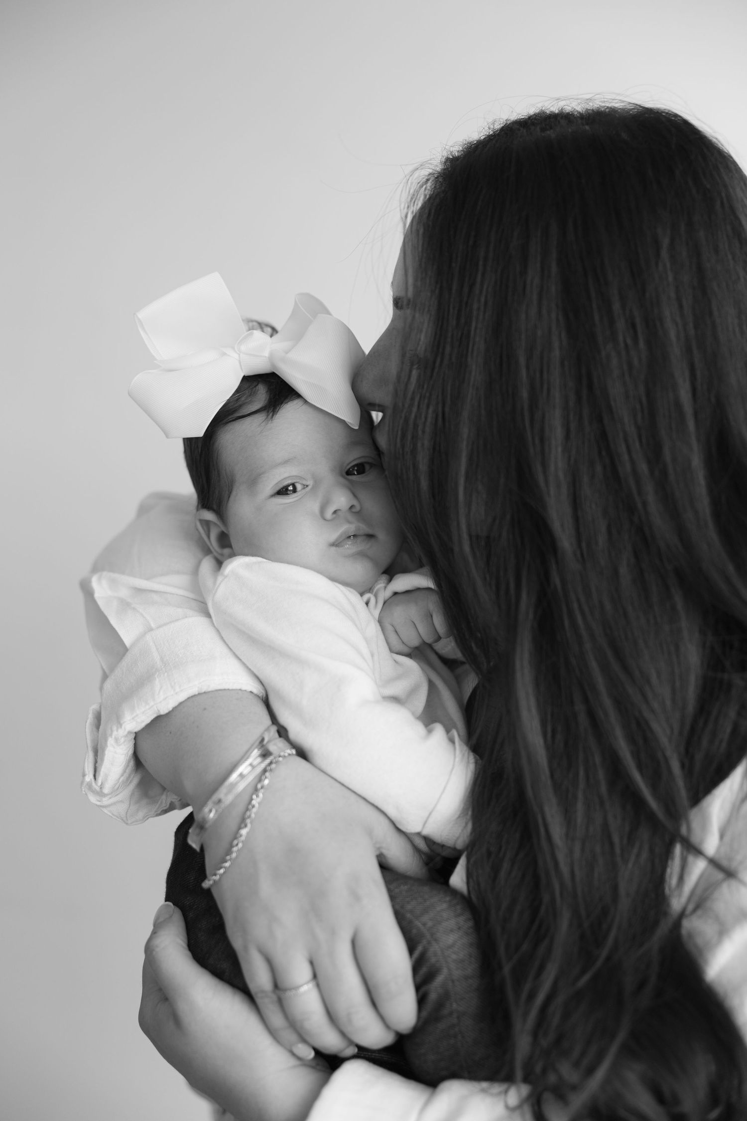 A black and white portrait of a small baby wearing a white bow being held by someone with long dark hair.