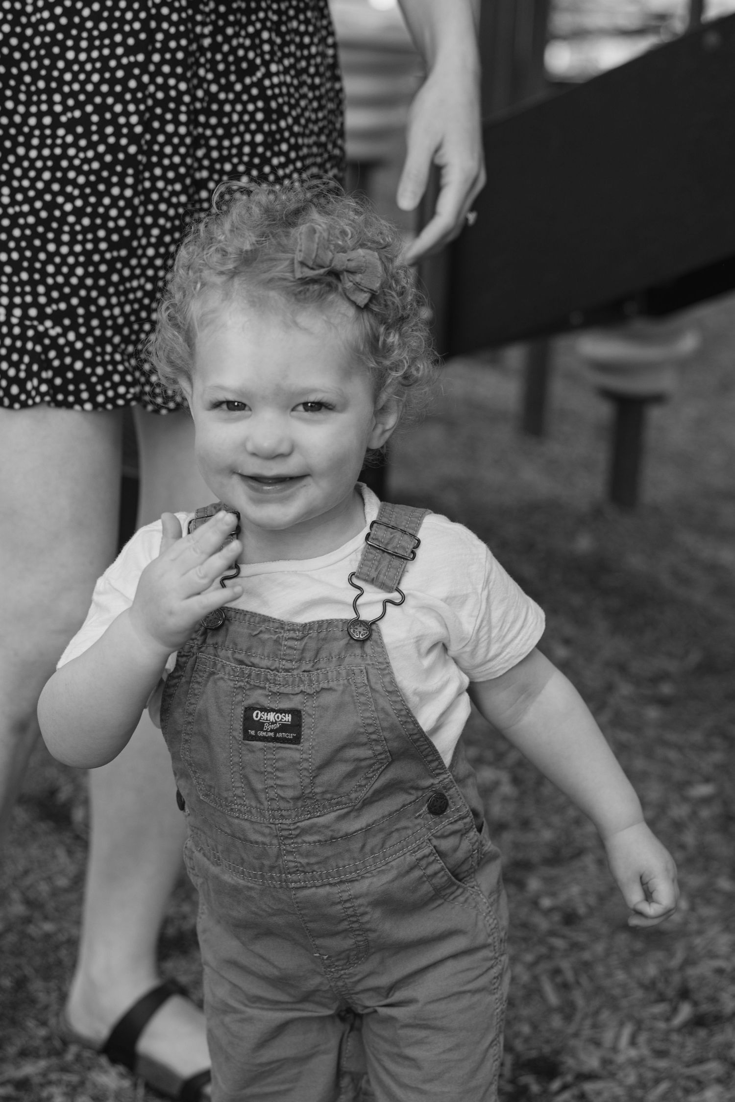 Black and white photo of small child in OshKosh overalls smiling with curly hair outdoors.