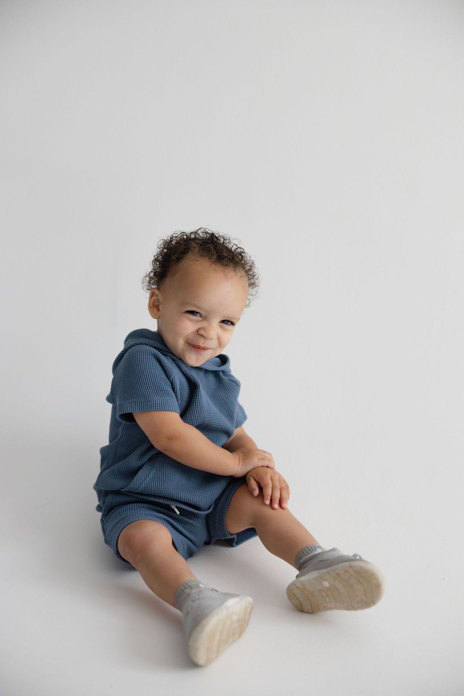 A small child in a blue outfit sits on a white background wearing gray sneakers.