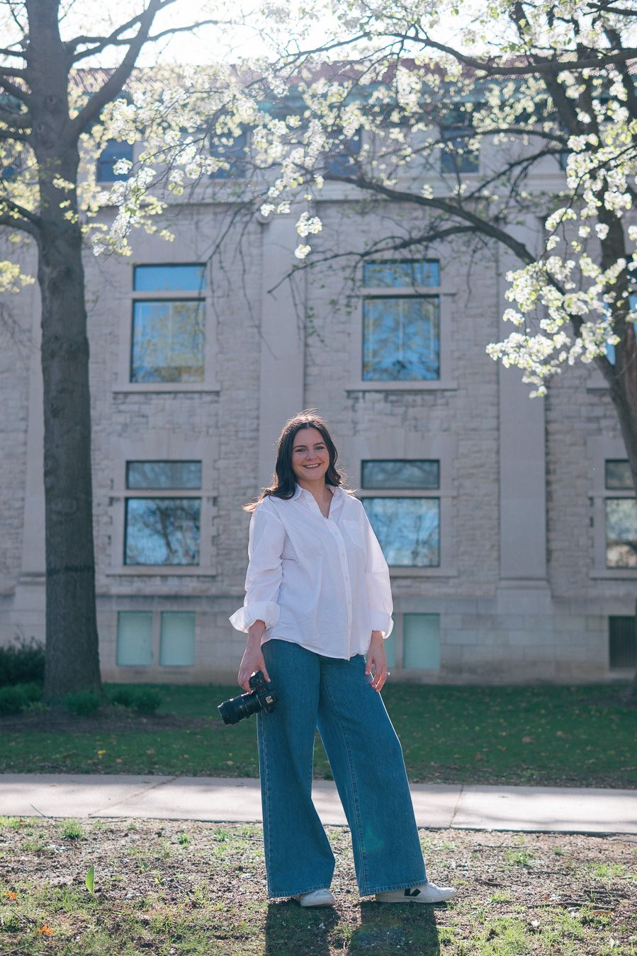 A person in a white blouse and blue jeans stands in front of a brick building with blooming spring trees.