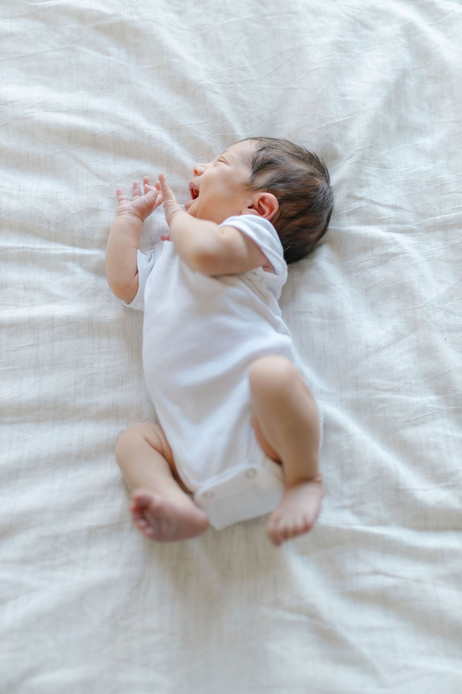 A baby in a white onesie yawns while lying on soft white bedding.