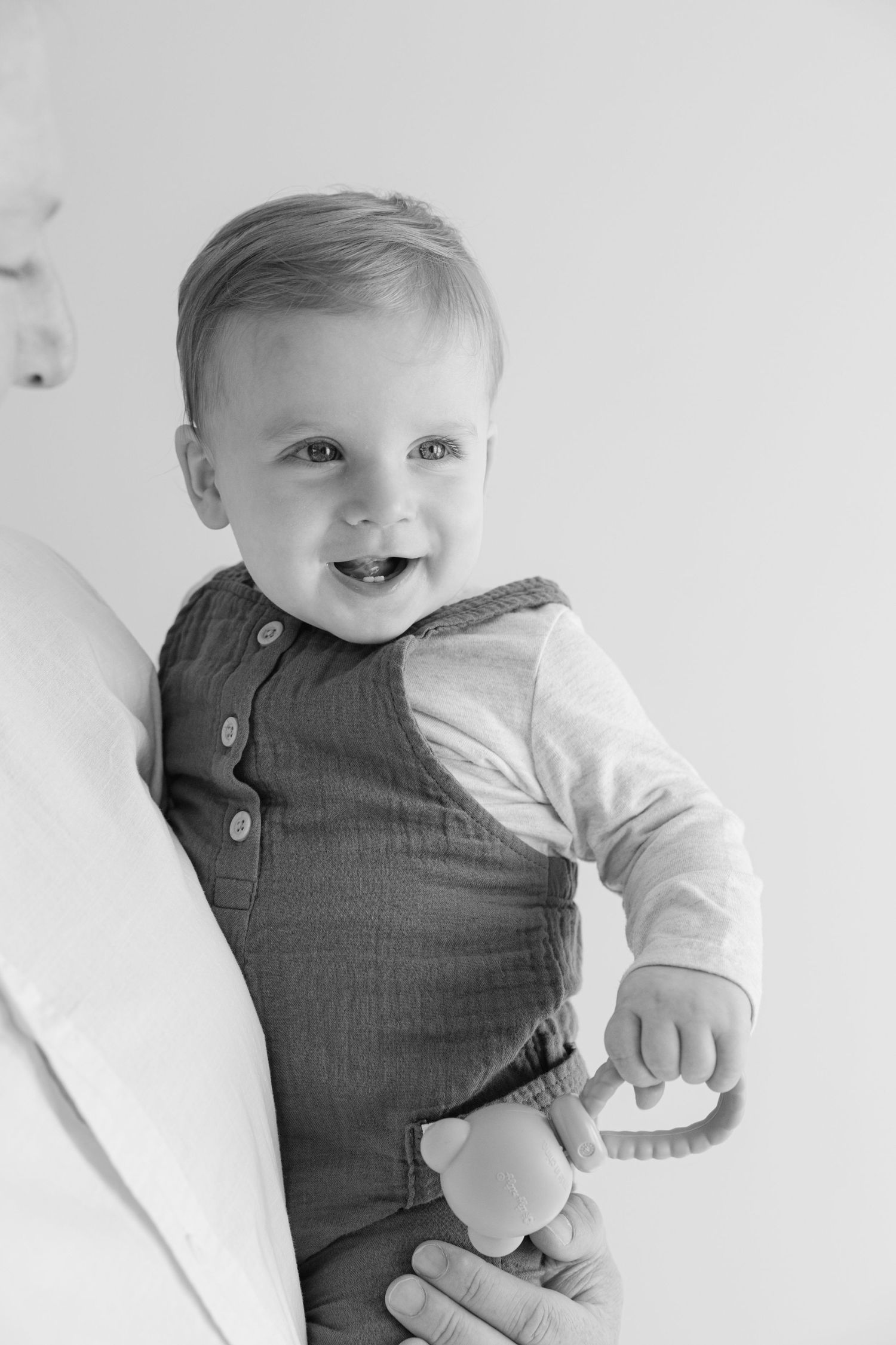 Black and white portrait of a smiling baby wearing a gray vest and white long sleeve shirt lying on a white surface.