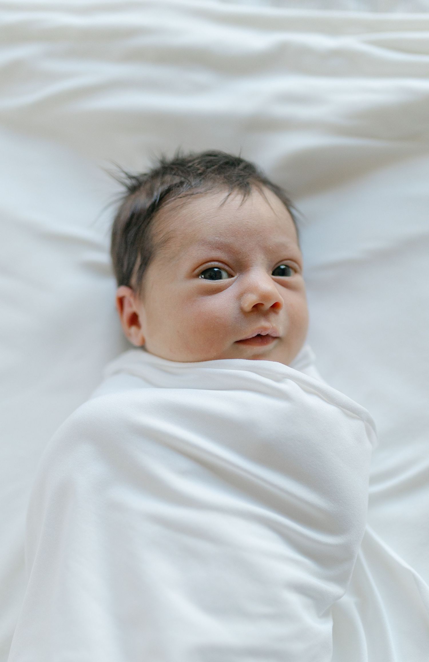 A newborn baby wrapped in soft white fabric lies on white bedding looking alert and content.