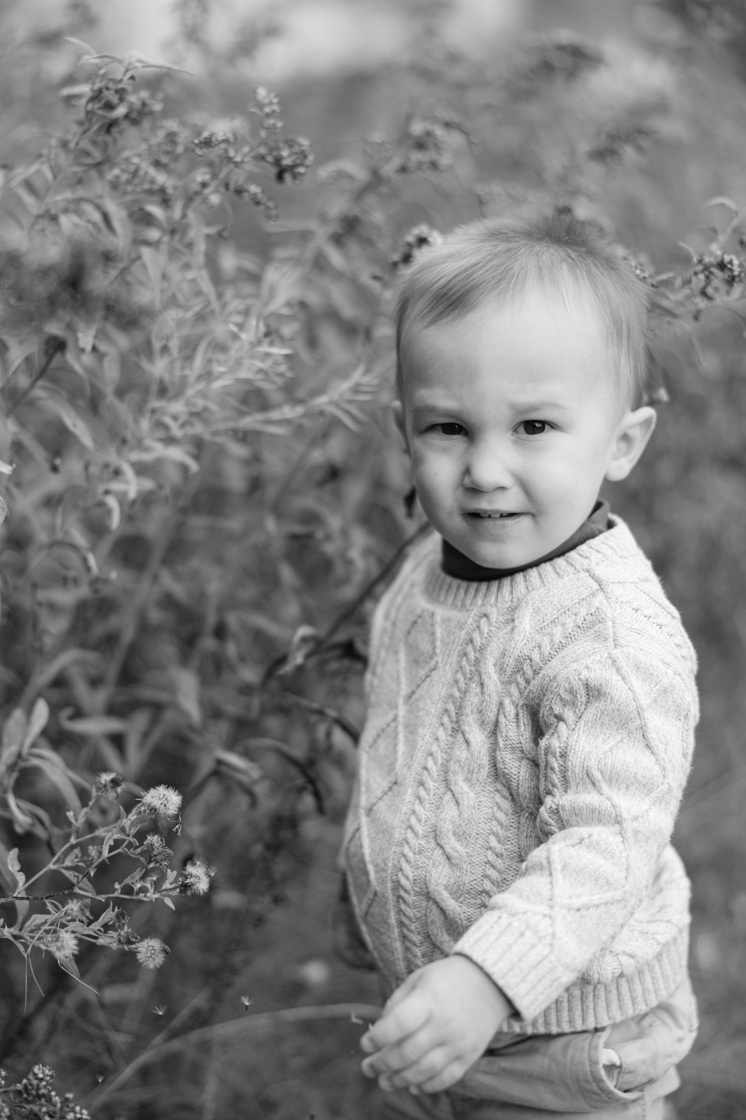A child in a white cable knit sweater poses outdoors against a natural background in black and white photography.