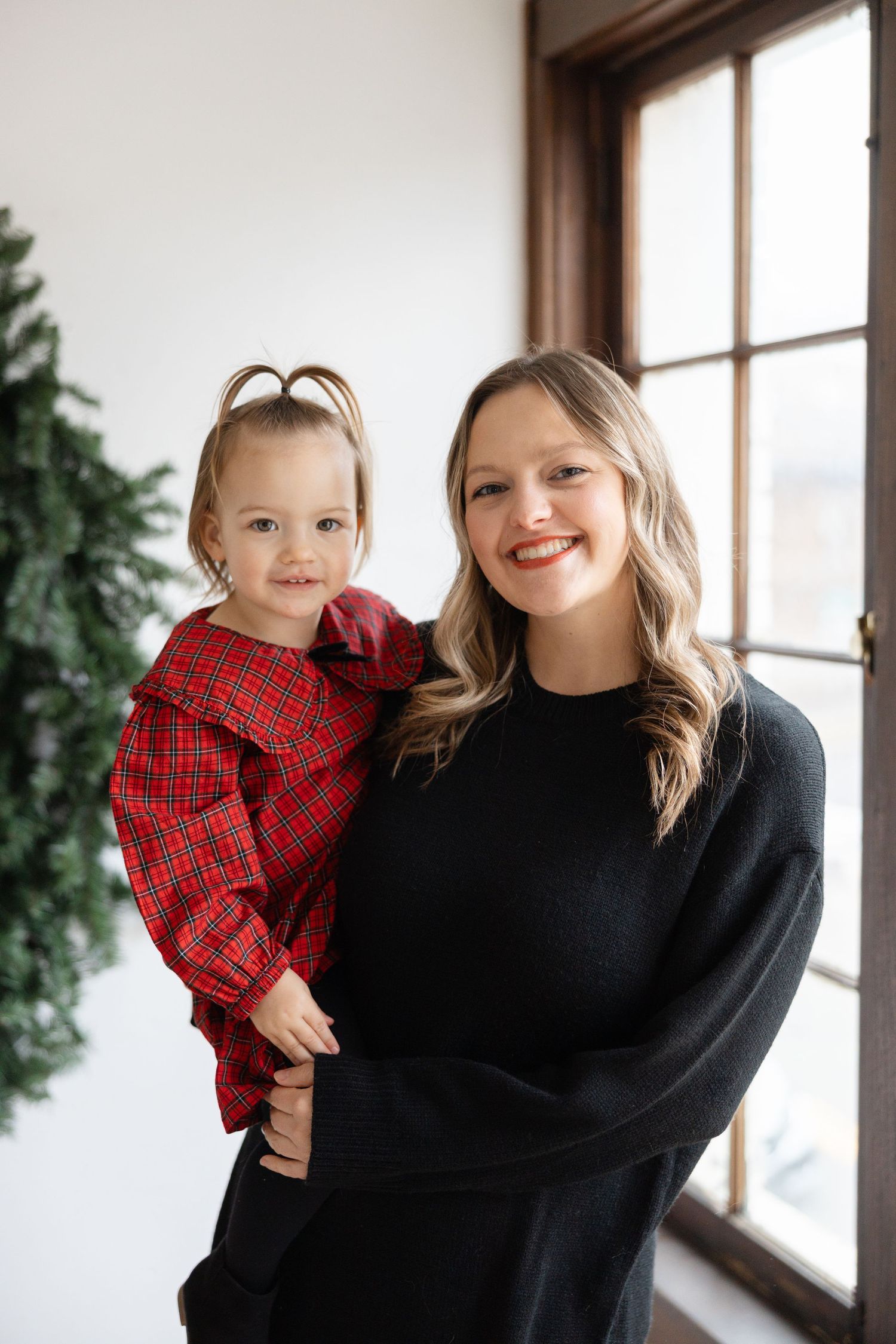 A parent in a black sweater holds a small child wearing a red plaid shirt in front of a bright window.