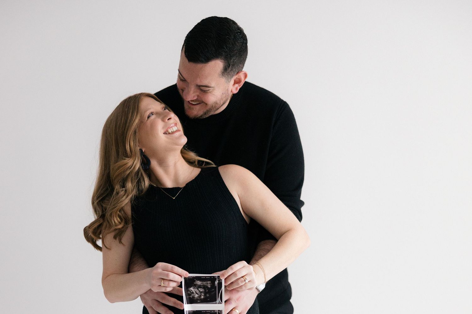 A couple in black clothing embraces while holding a sonogram photo against a light gray background.