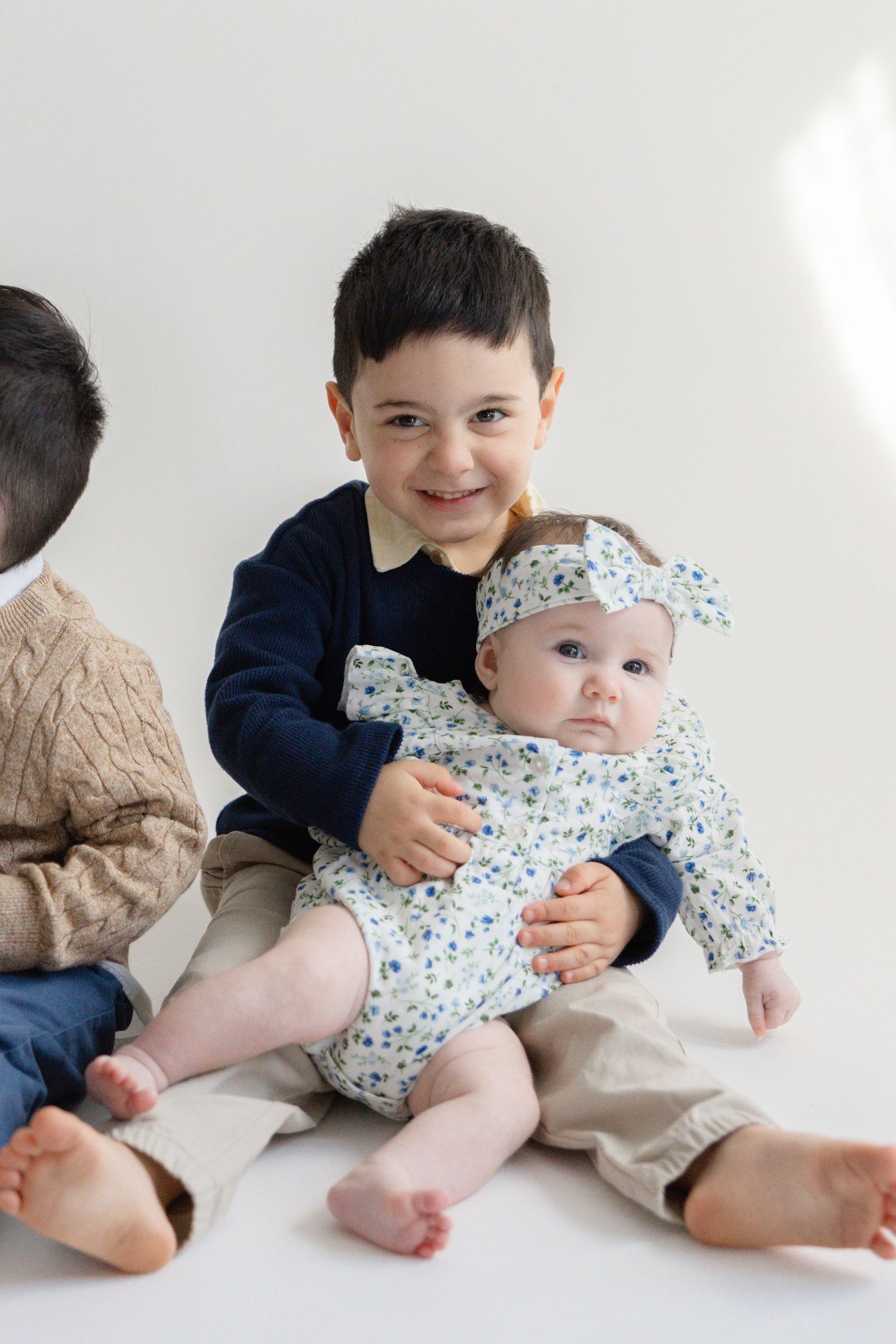 Two siblings share a tender moment as the older one gently embraces and interacts with the younger baby on a white backdrop.