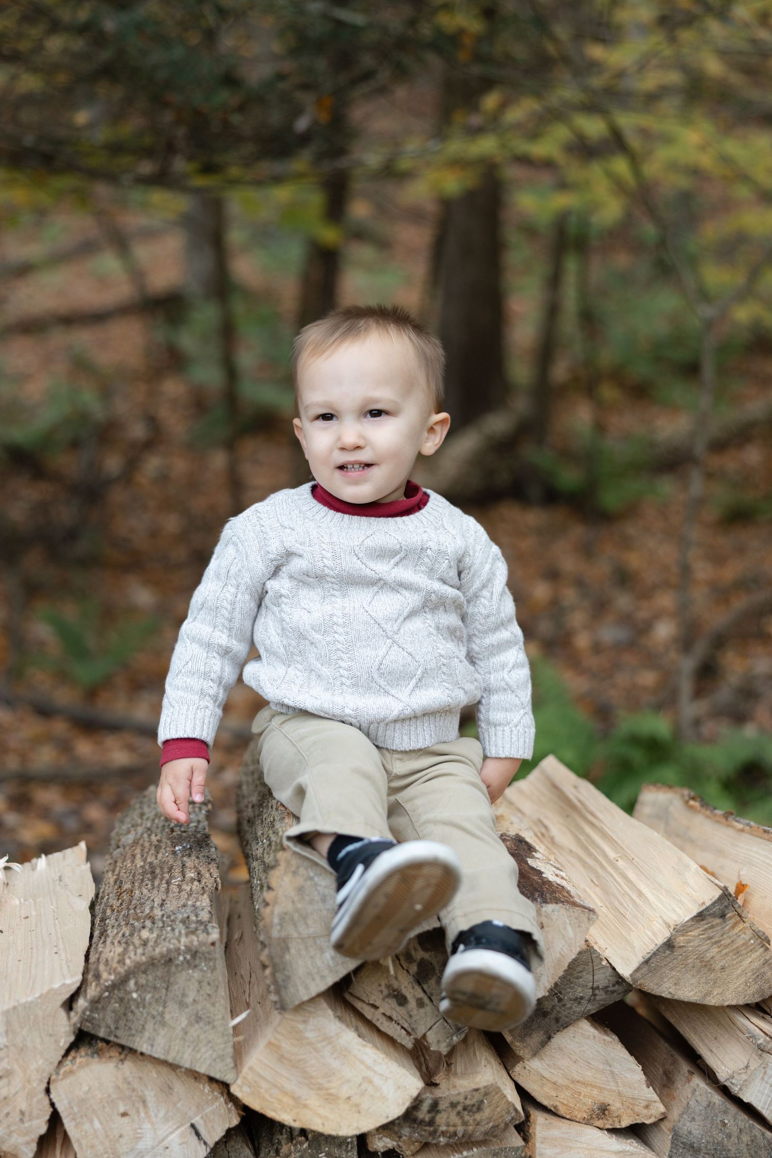 A small child in a white knit sweater sits playfully on a stack of chopped firewood in an autumn setting.
