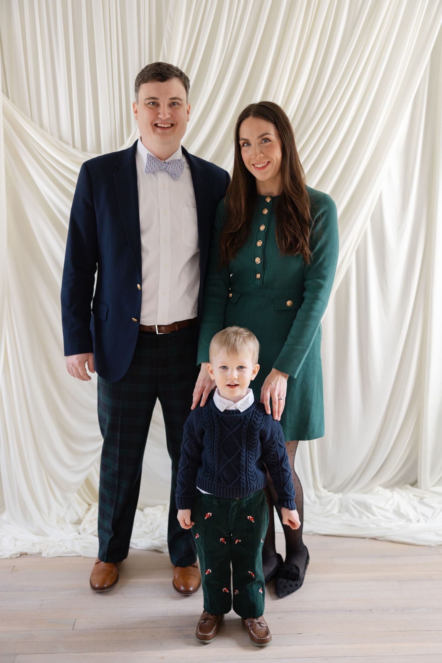 Family portrait series against white draped backdrop showing parents and toddler in coordinating navy and green outfits.