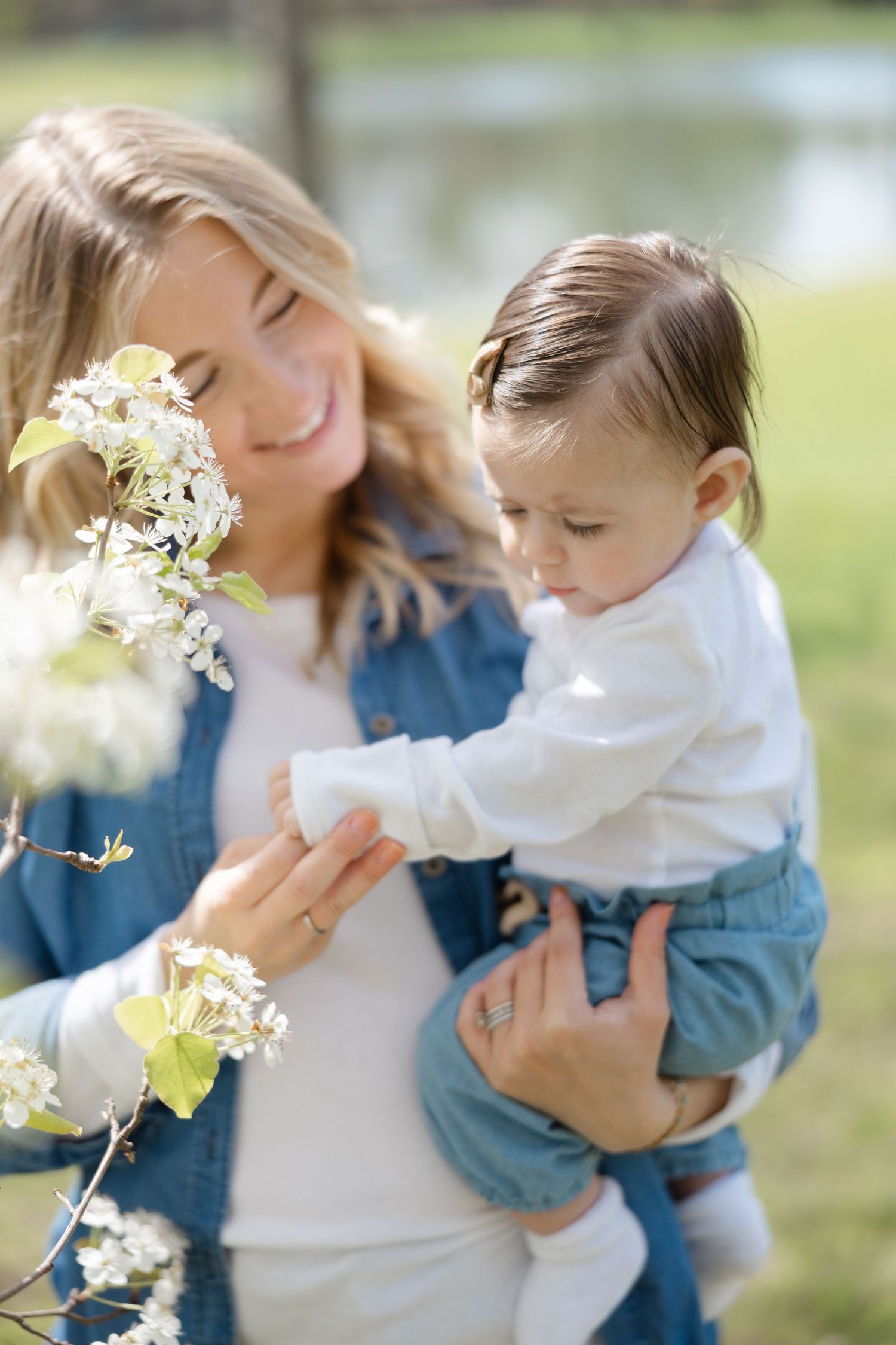 A smiling mother in denim holds her baby near white spring blossoms in a sunny park.
