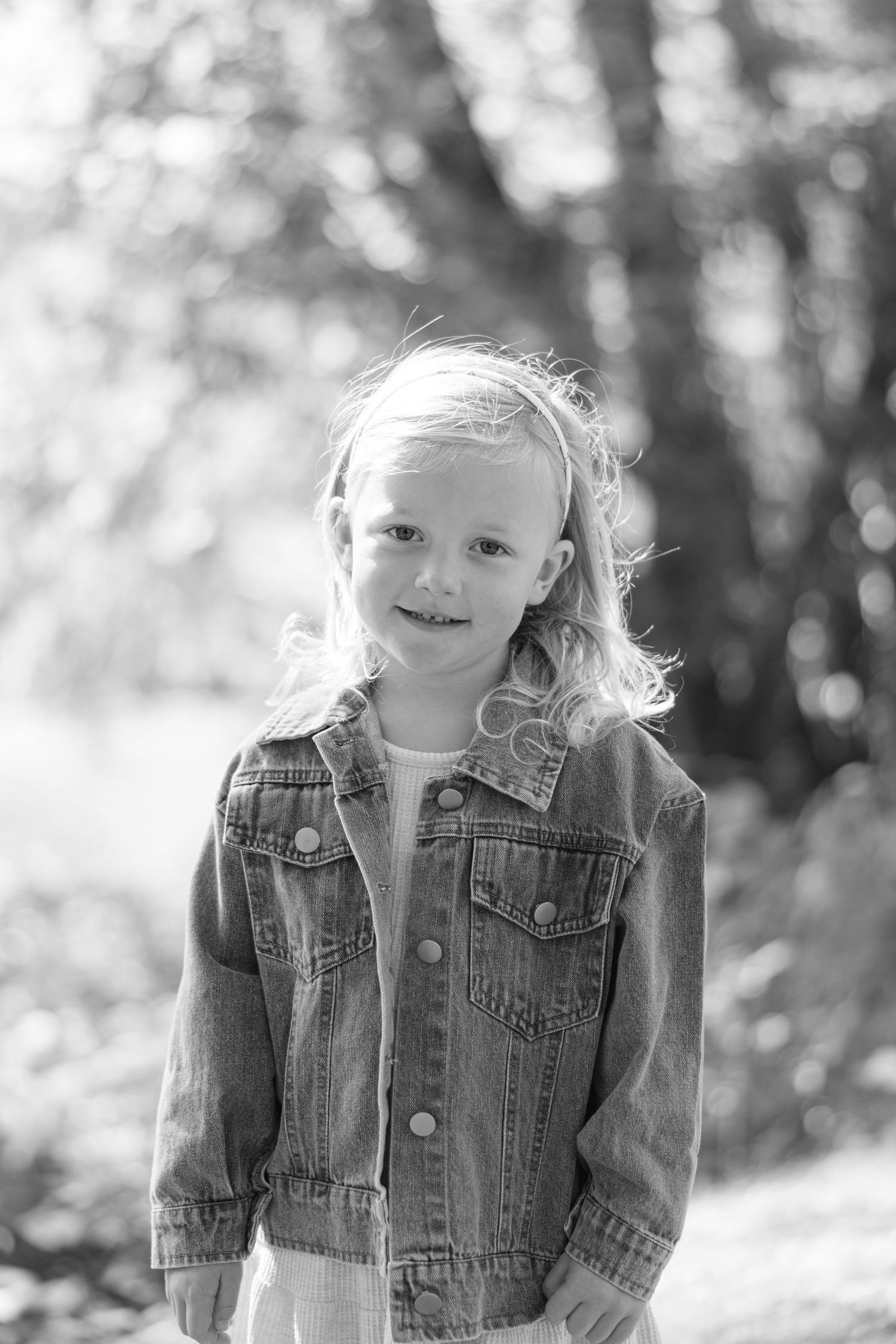 Black and white portrait of a child wearing a denim jacket and smiling outdoors in a natural setting.