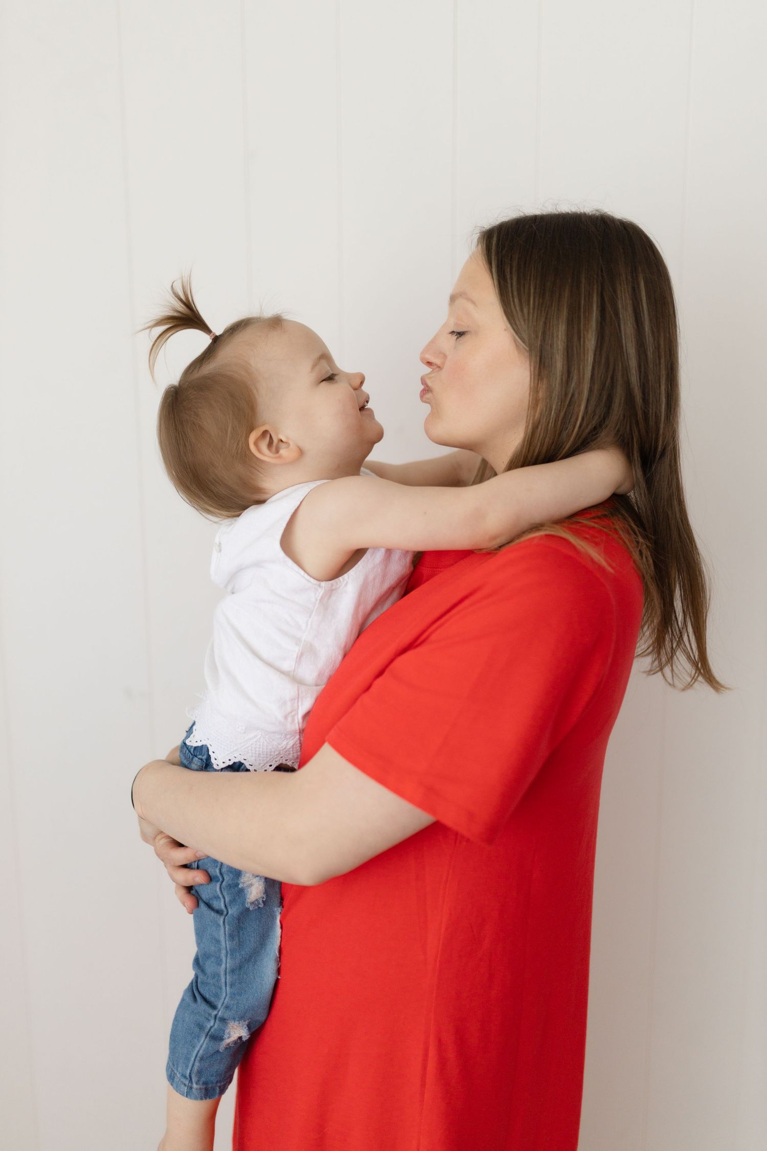 A parent in a red shirt shares a joyful moment with a baby in white against a neutral background.