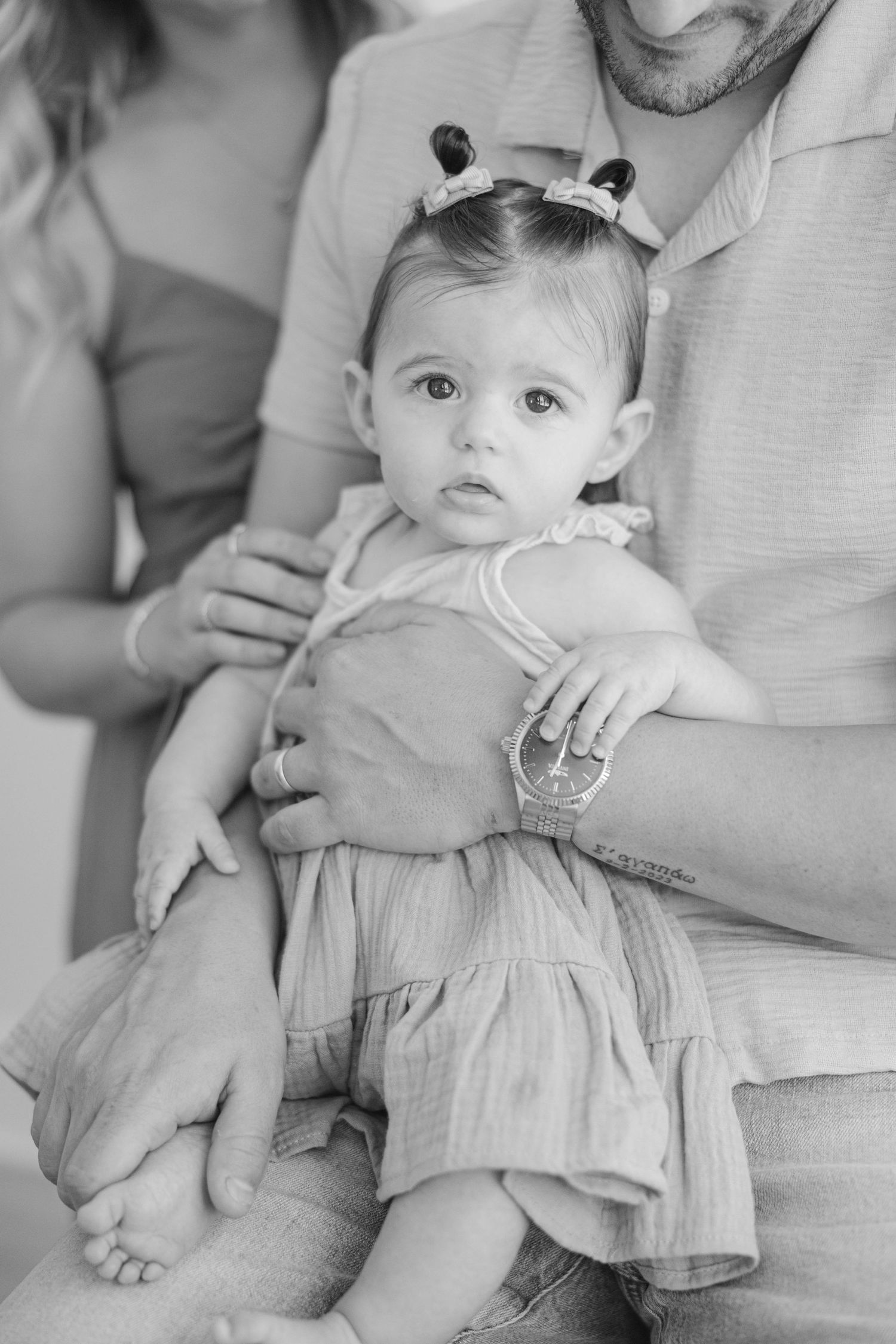 Black and white photo of a baby girl in a ruffled dress and headband being held by her parents.
