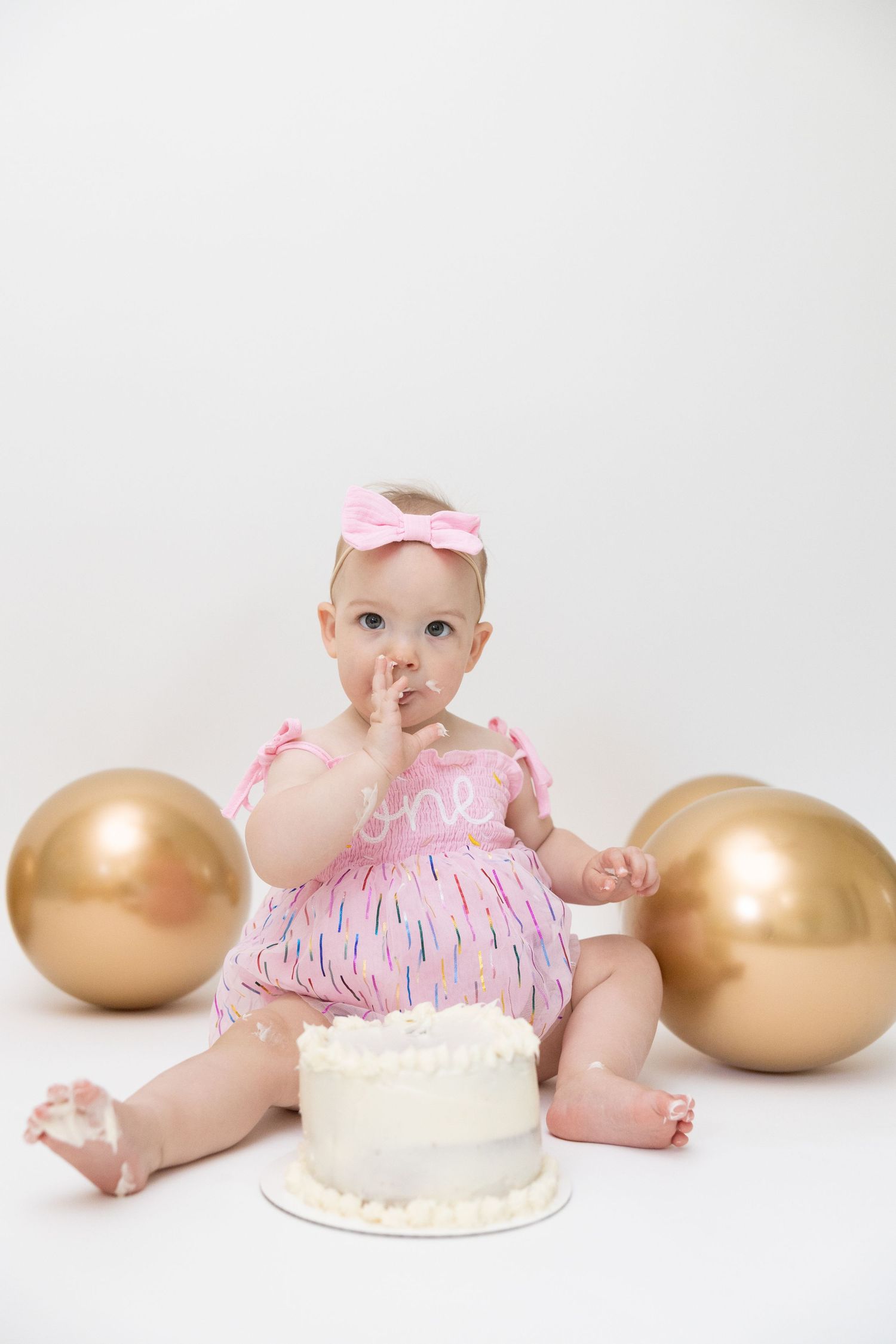 Baby in pink dress sits with white cake and gold balloons during first birthday cake smash photo session.