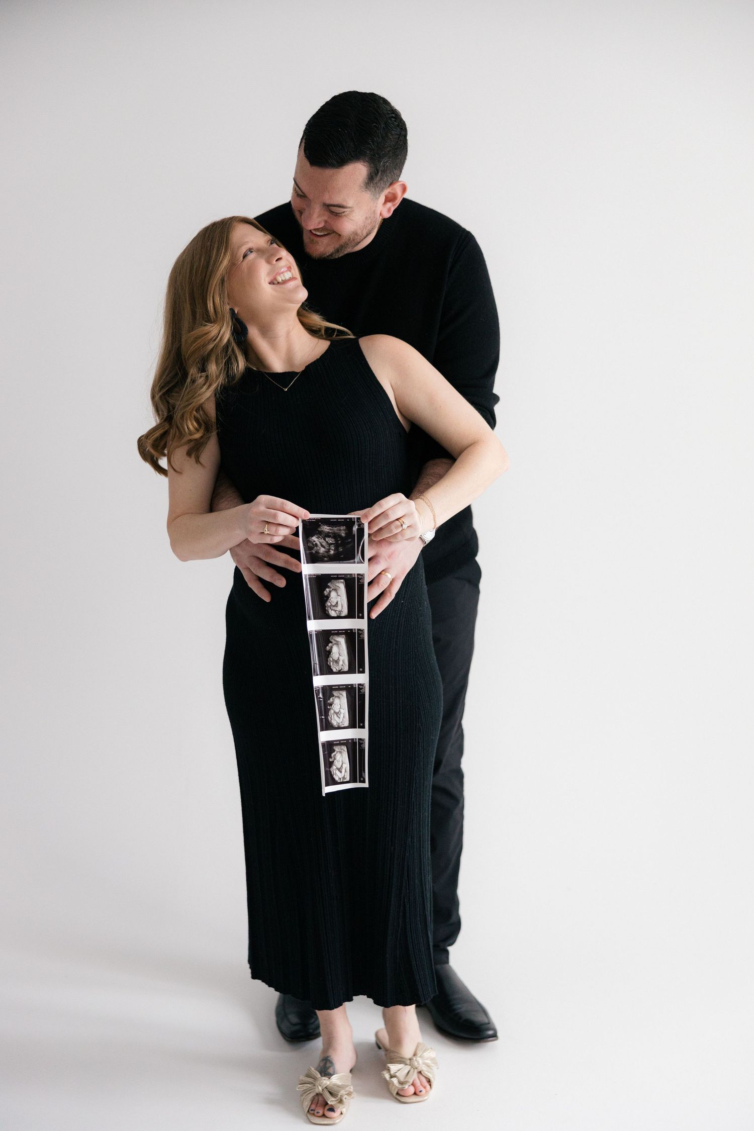 A couple in black outfits share a joyful moment while holding ultrasound photos against a white backdrop during a maternity photoshoot