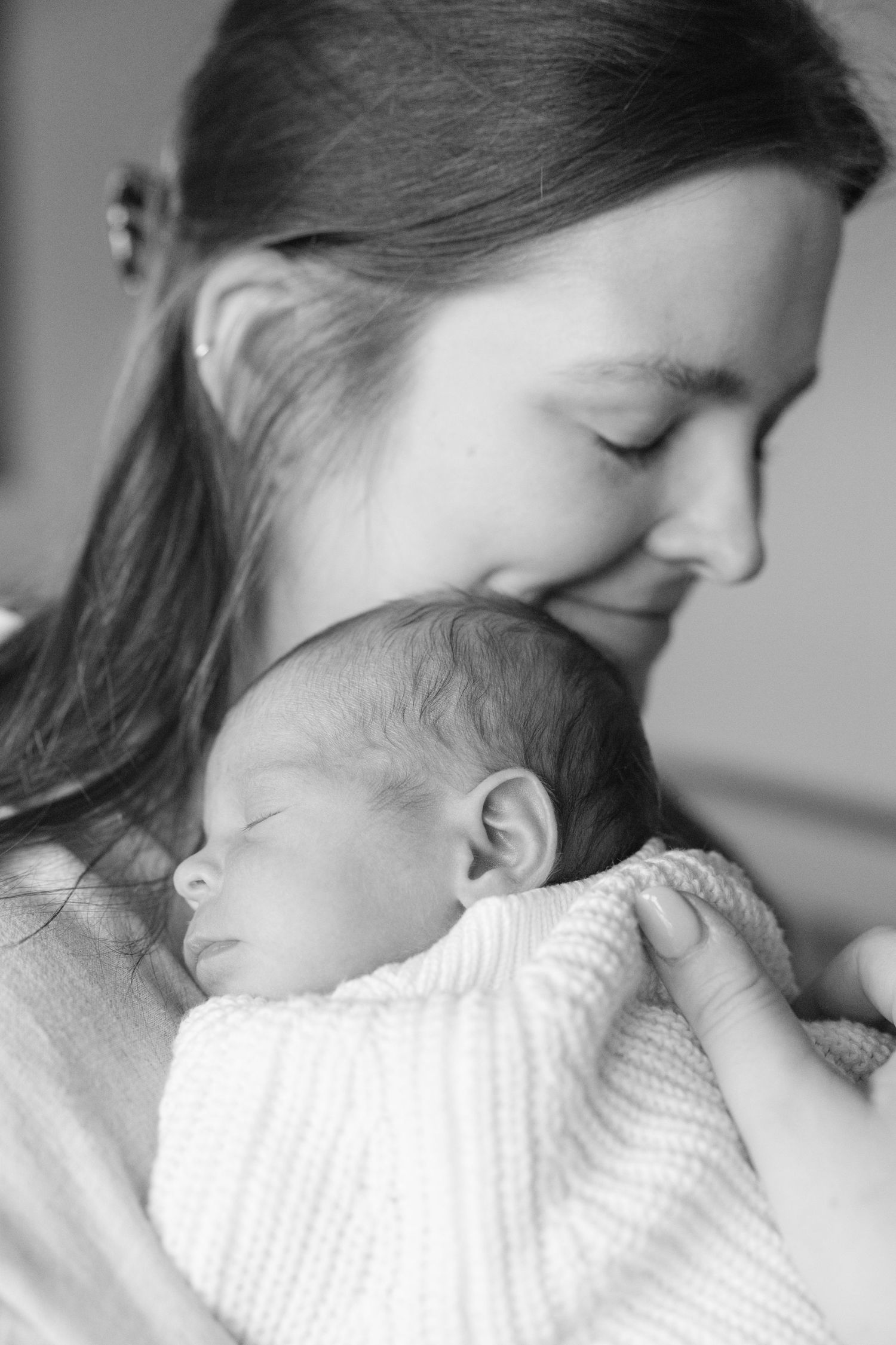 Black and white photo of a smiling mother tenderly holding her sleeping newborn baby wrapped in a white blanket.