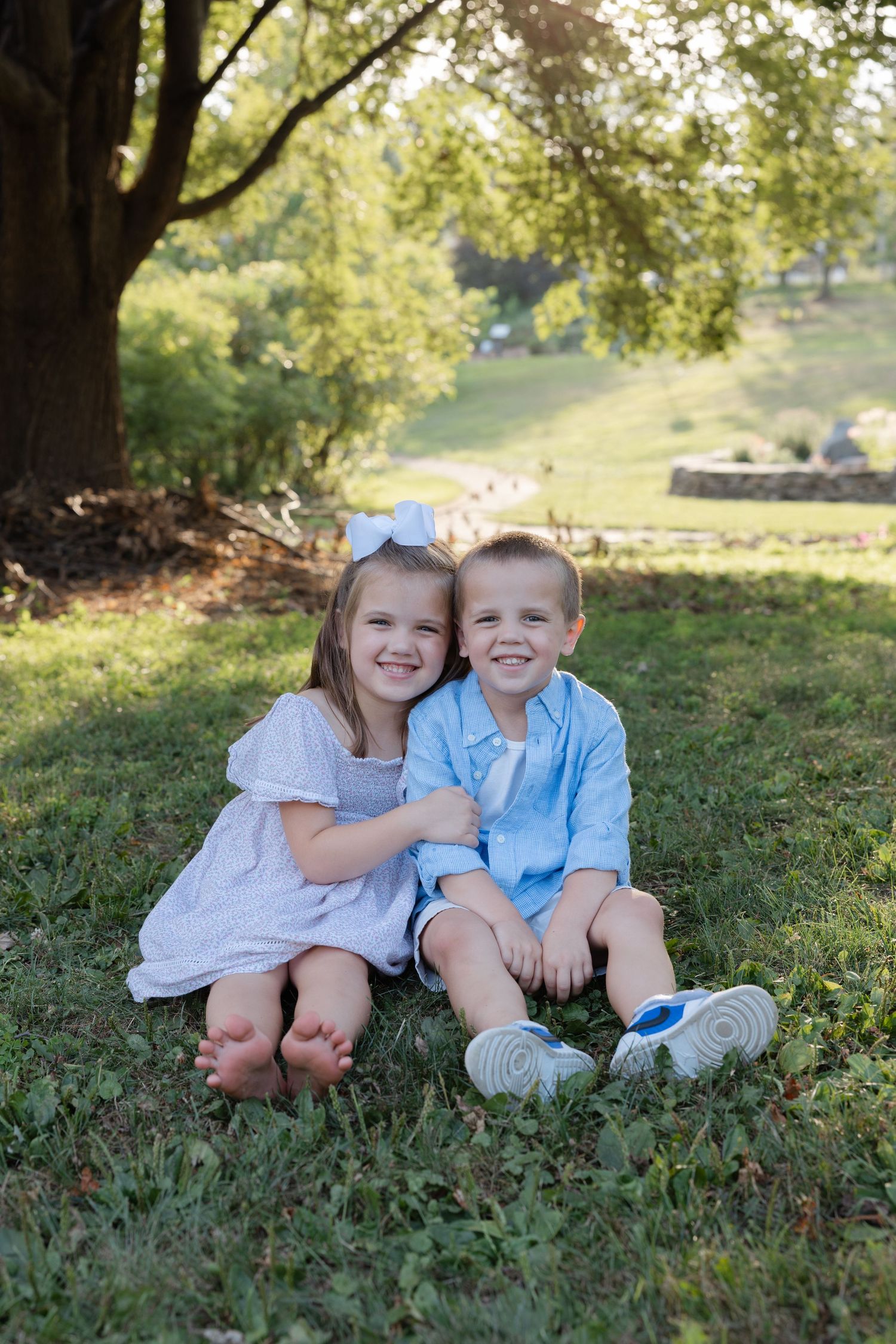 Two children sit close together on grassy ground under dappled sunlight in a park setting.
