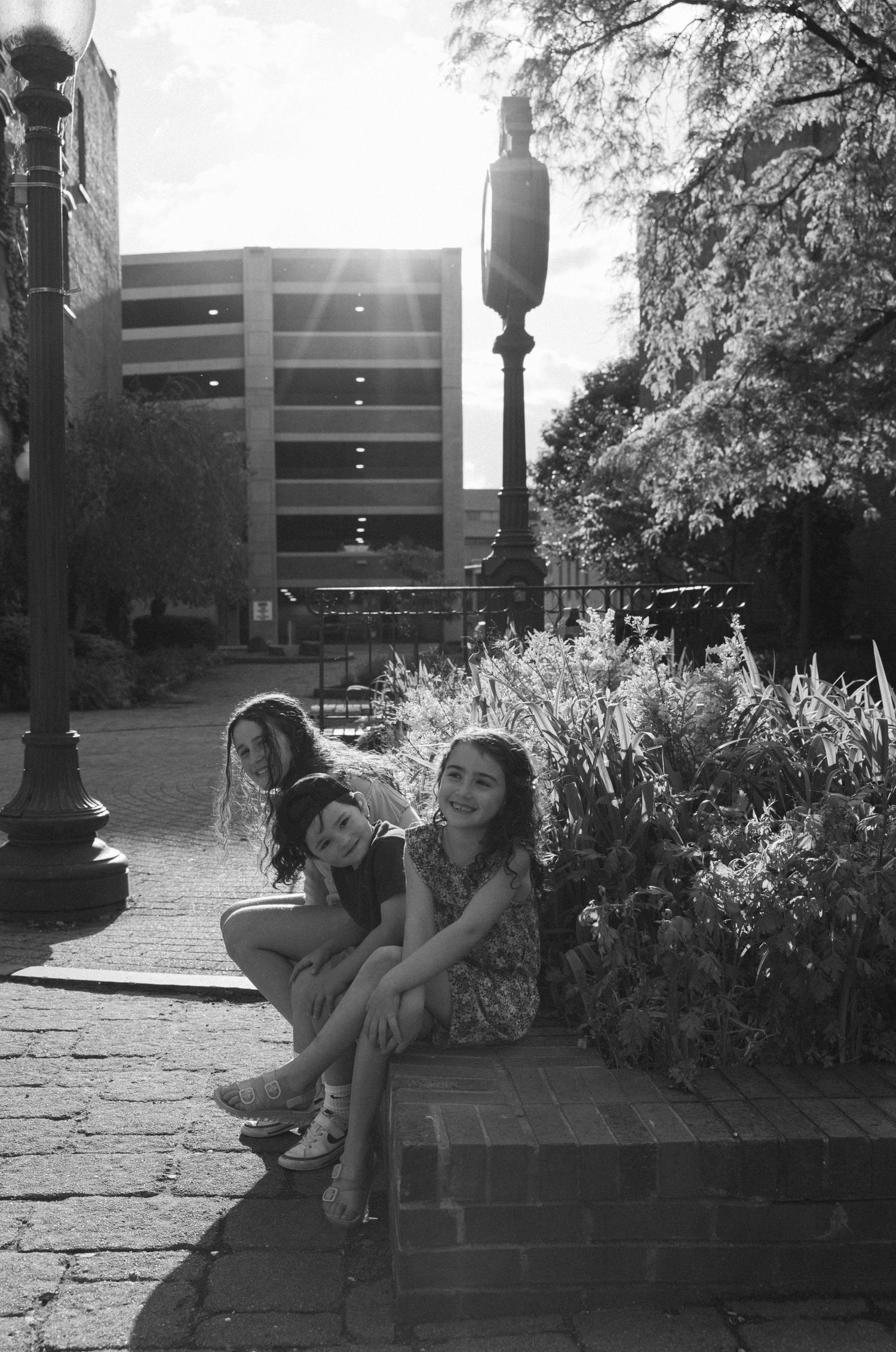 Two people sitting on brick steps next to lamp post and garden on a sunny urban plaza.