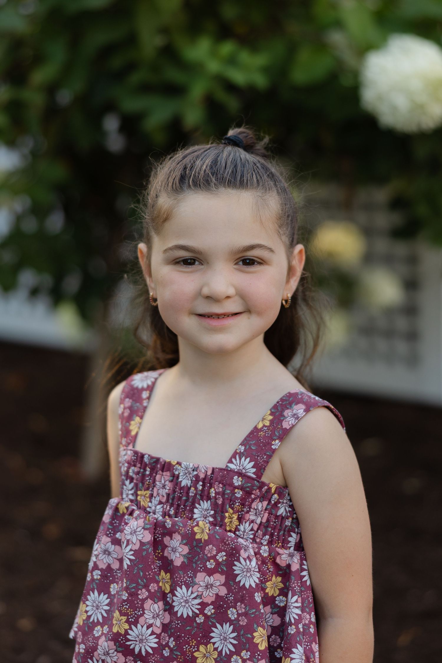 A young child in a purple floral sundress smiles while posing outdoors against a natural background.