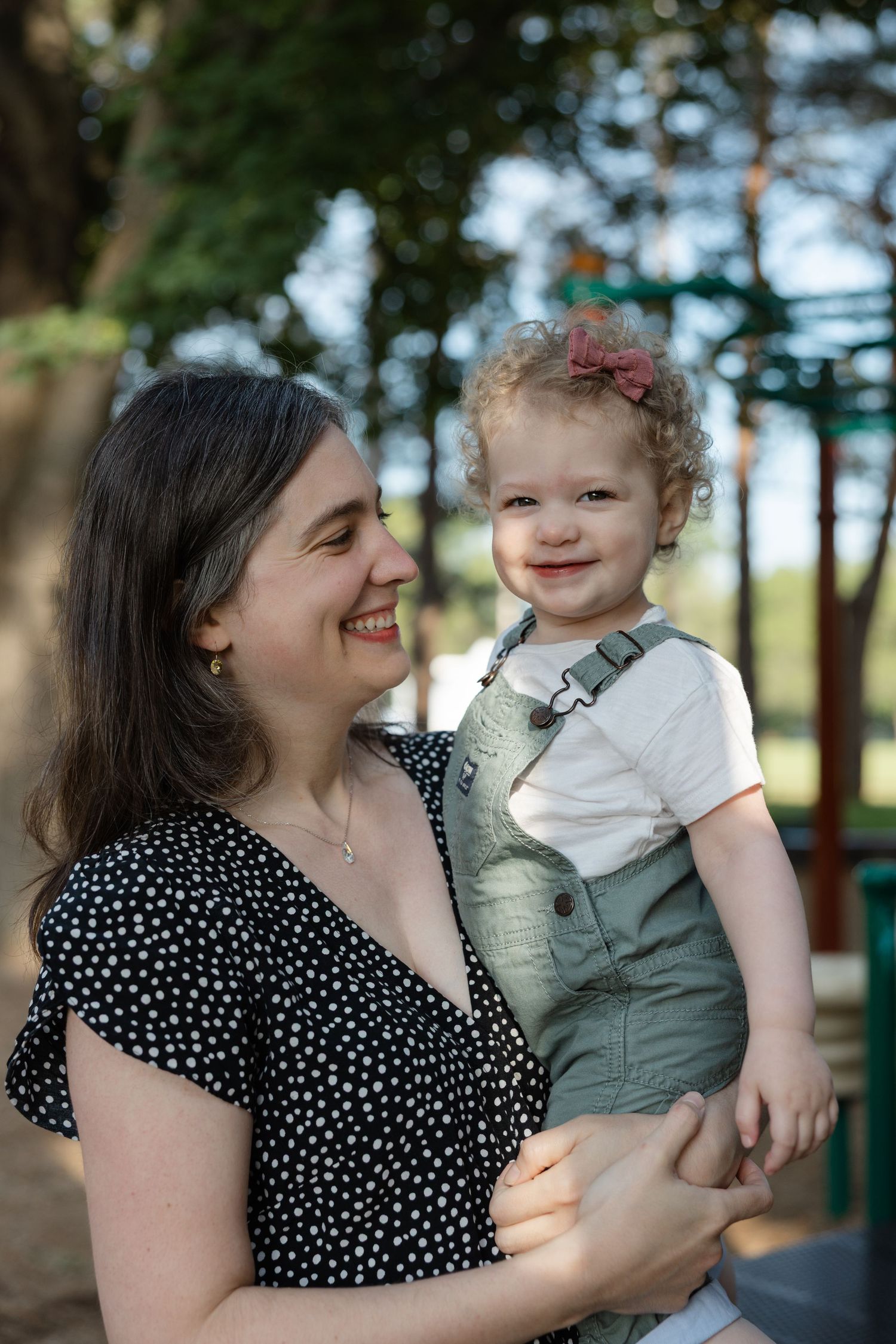 A parent in a polka dot dress holds a toddler wearing mint green overalls and a white shirt outdoors on a sunny day.