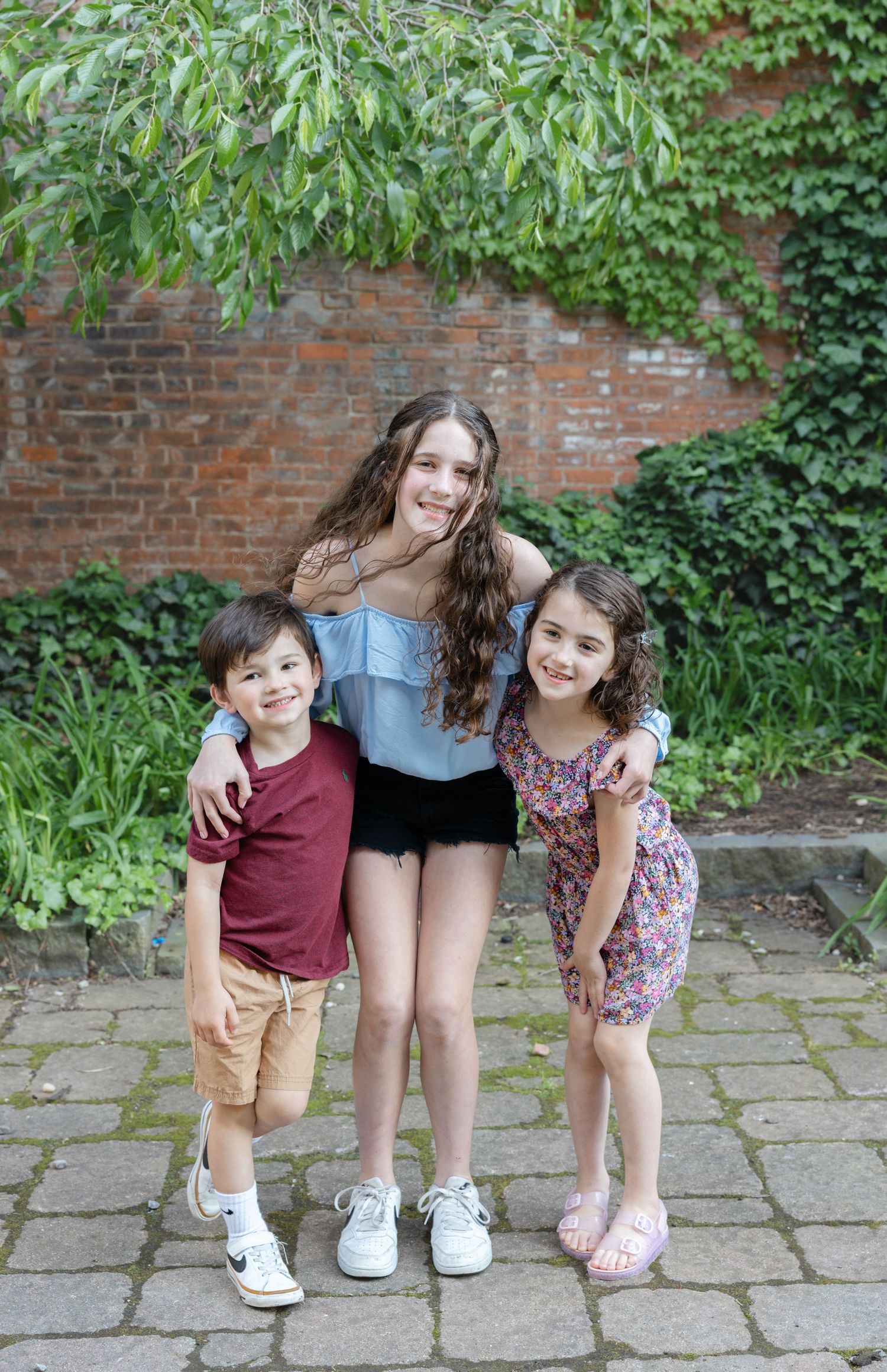 Three children in casual attire pose together on stone pavement in front of a brick wall with green foliage.