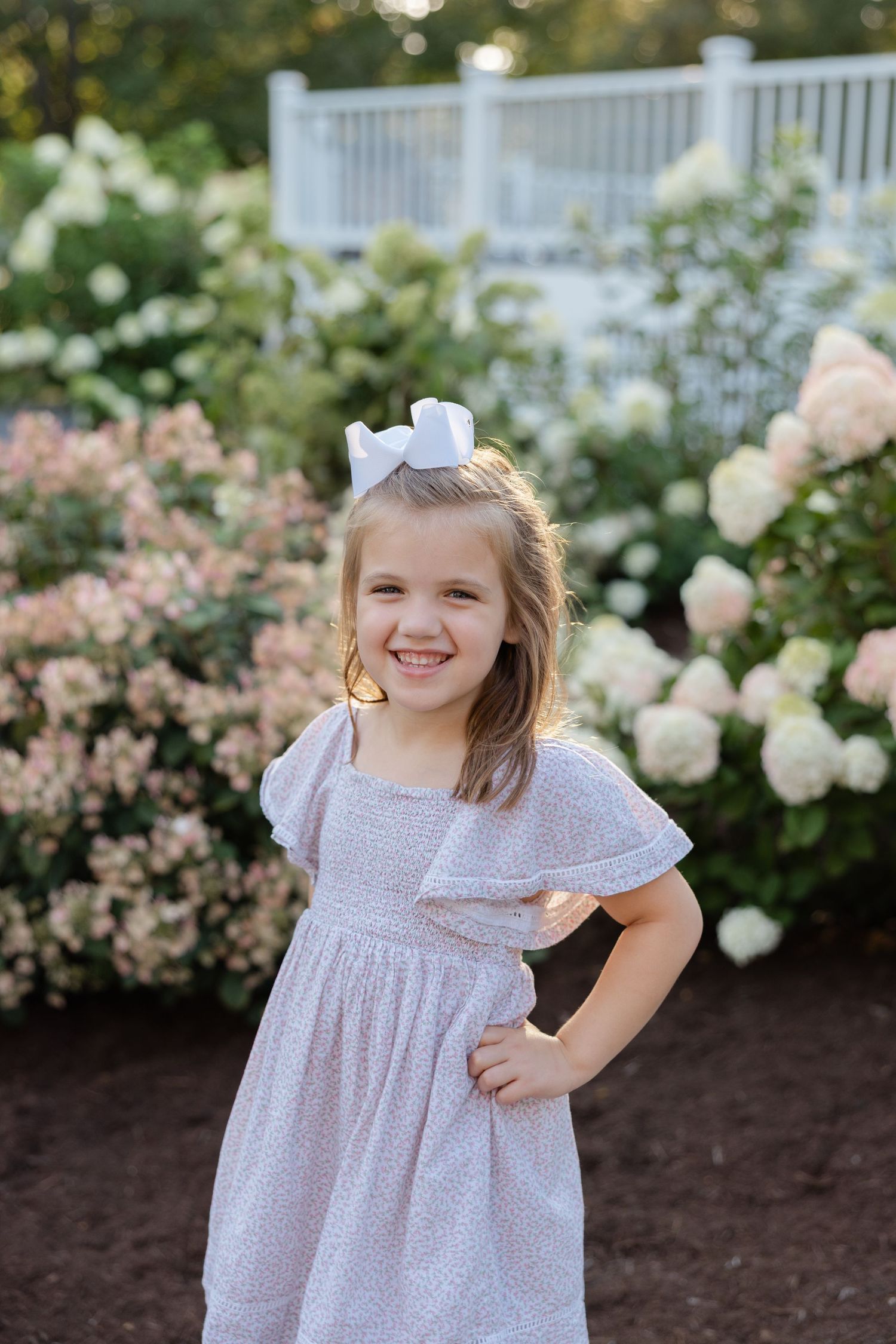A young person wearing a white and purple floral dress poses in a garden with white and pink flowers in the background.