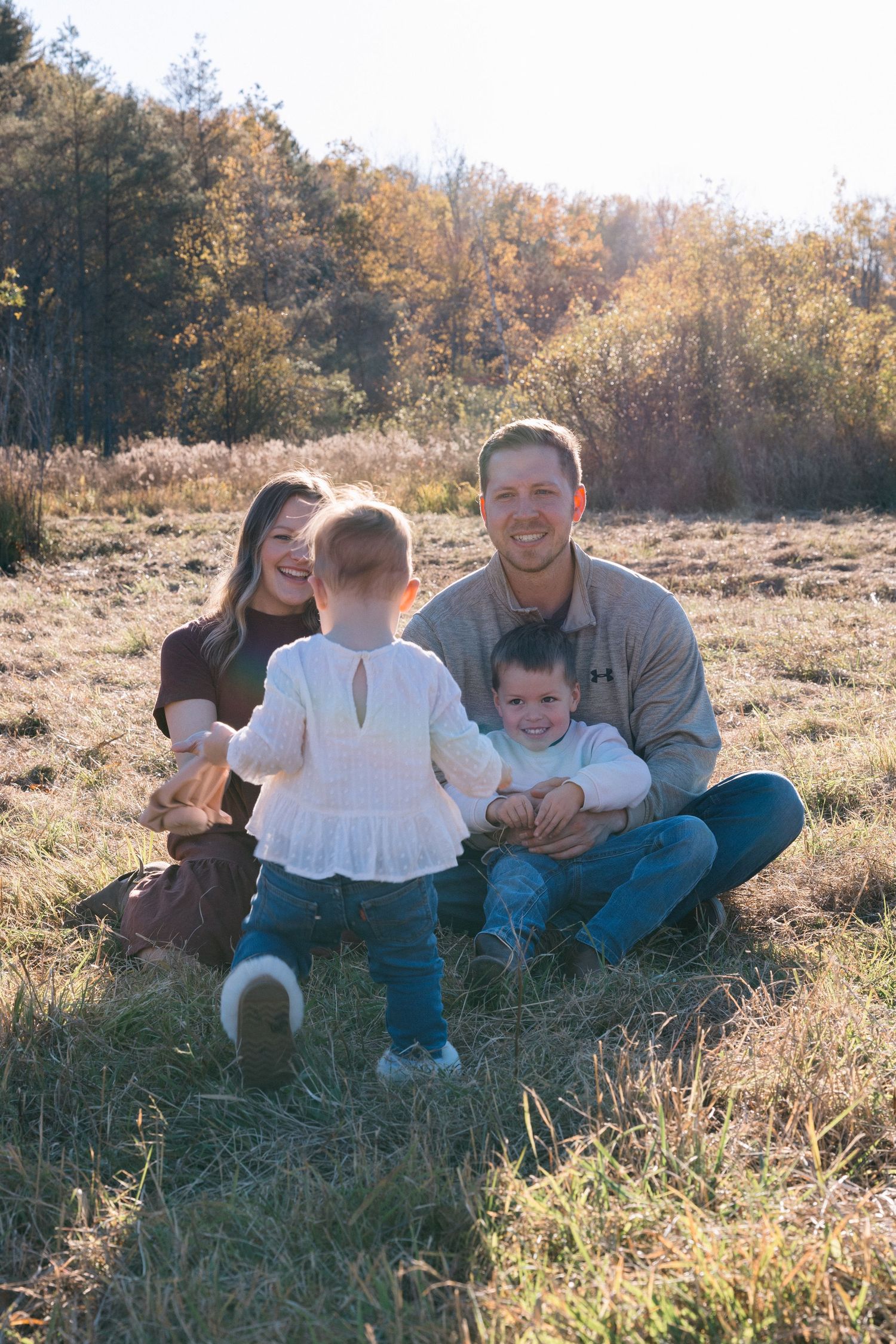 A family sits together on grassy fall field with autumn trees in the background during golden hour.