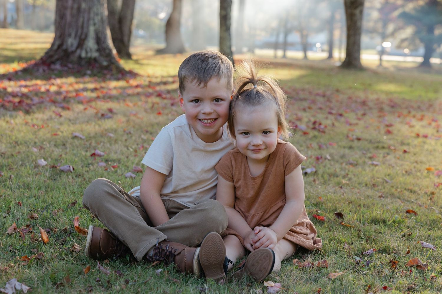 Two children sit together on autumn grass with fallen leaves and trees in a sunlit park setting.